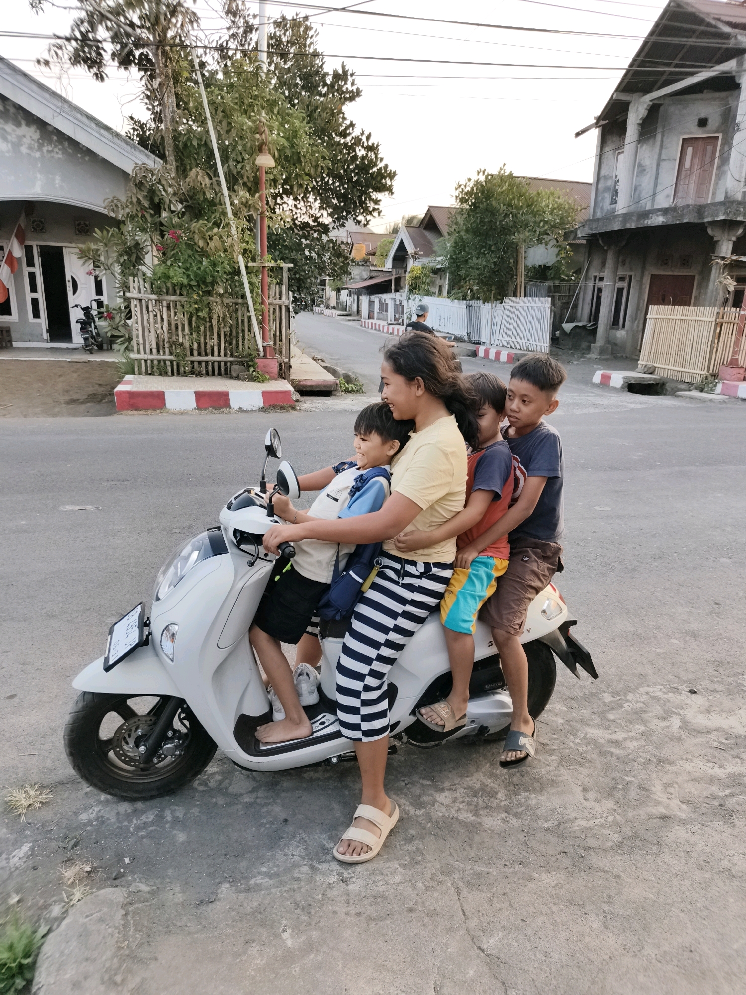 In Tombatu, Indonesia, a woman is driving a white scooter with four children riding on it. The woman is in the front, and three children are behind her. The scooter is on a paved road. In the background, there are houses and trees. The sky is overcast.