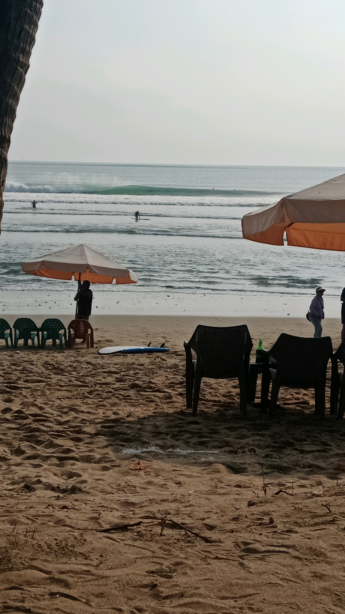 The image captures a serene beach scene in Kuta, Indonesia, with the ocean and sky dominating the background. In the middle ground, several people are visible on surfboards in the water, engaging in surfing activities. The sandy shore features beach chairs and umbrellas, with a few people leisurely walking along the beach. A surfboard rests on the sand near the foreground. The overall atmosphere is tranquil, with soft lighting suggesting it might be early morning or late afternoon. The presence of surfboards and the calm waves indicate a popular spot for surfing.