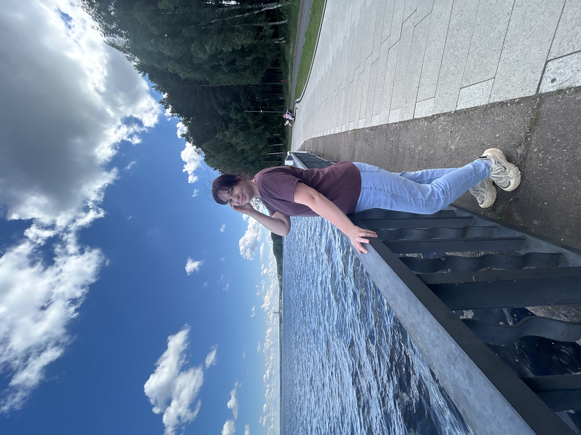 A young woman with short, dark hair is lying down on a dark-colored metal structure, which appears to be a set of stairs leading down to the water. She is wearing a maroon t-shirt and light blue jeans, and white sneakers. Her left arm is extended forward, resting her head on her hand, and her right arm is bent, with her hand resting on the railing. She is looking towards the camera with a slight smile, suggesting a relaxed and casual mood.
The scene is set by a large body of water, likely a river or lake, with gentle ripples on its surface. The sky is a vibrant blue with scattered white clouds, indicating fair weather. Behind the woman, a paved walkway runs alongside the water, bordered by a line of trees. In the distance, a lighthouse-like structure can be seen on the horizon. The overall impression is a pleasant day outdoors, possibly in a park or recreational area, given the paved walkway and presence of trees. The time of day appears to be daytime, judging by the bright sunlight and blue sky. No visible text is present in the image. The location context suggests this scene is in Chaikovsky, Russia.