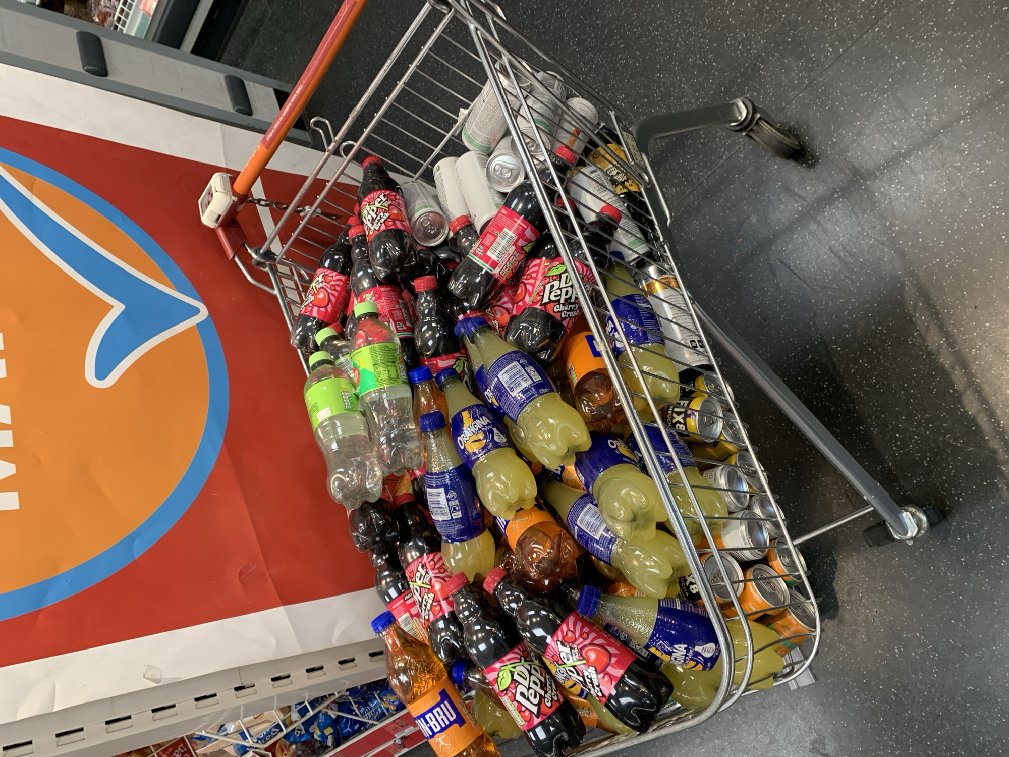 A wire shopping cart, mostly full of soda and cans, sits in an indoor setting, presumably a store aisle. The cart is angled and positioned near a display with orange and blue markings, potentially advertising the store or a product. The items inside include bottles and cans of various brands, such as "IRN-BRU", "Dr Pepper Cherry Crush", "Orangina". Cans are also visible. The floor is dark gray with light-colored speckles. The cart handle extends towards the upper right.