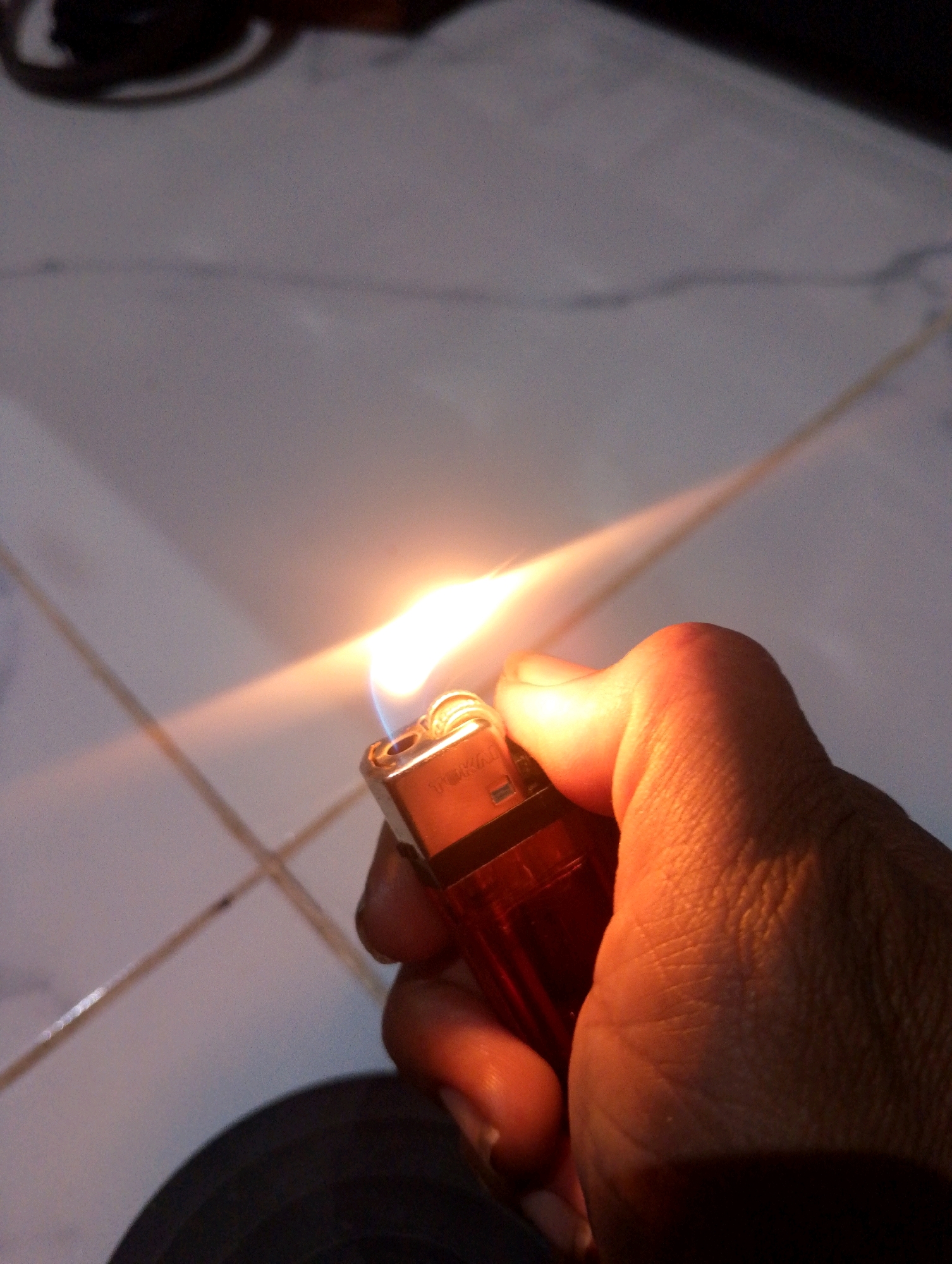 In a close-up, low-angle shot, a hand is holding a lit red and gold lighter. The flame from the lighter is bright orange and yellow, with a hint of blue at the base. The hand, which appears to be that of an adult, is illuminated by the light of the flame, casting shadows on the textured skin. The background is a tiled floor, with visible grout lines. The lighting suggests it might be evening or indoors with artificial light, creating a warm glow. The scene is static, with no apparent movement. The location is specified as Sumbergirang, Indonesia.
