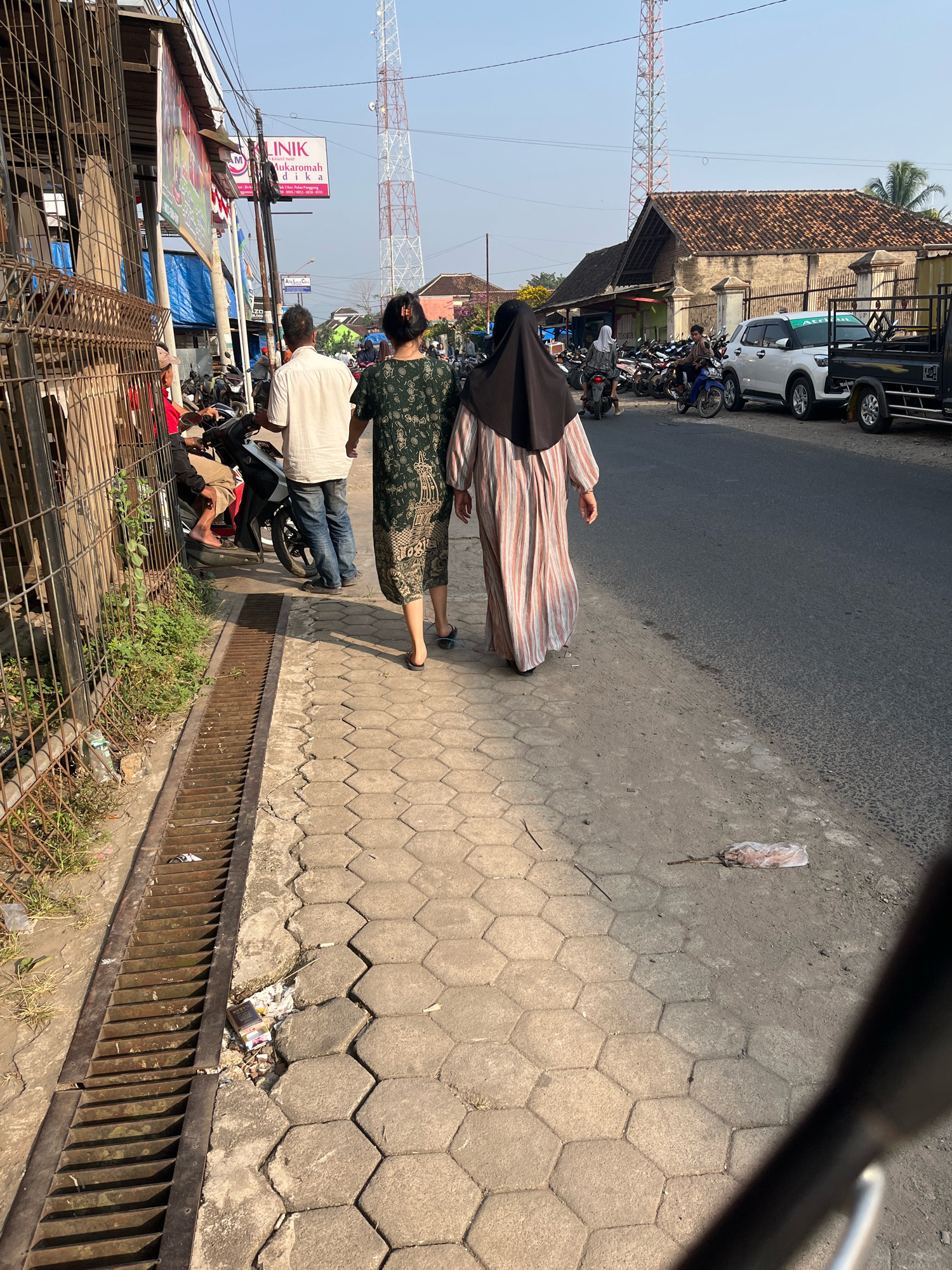 The image depicts a street scene in Kemuning, Indonesia, under a clear sky. Two individuals are walking away from the camera on a hexagonal-tiled sidewalk. The person on the right wears a dark hijab and a long, vertically striped dress in shades of pink and white. The person on the left, walking slightly ahead, wears a dark green patterned dress. To their left, a metal fence encloses some greenery and objects. A rusty metal grate runs along the edge of the sidewalk, bordering the street.
The street itself is paved and shows some wear. Several motorcycles are parked along the side, and a white car and a black truck are visible further down. Buildings with tiled roofs line the opposite side of the street, with a prominent cellular tower in the background. Signs are visible on some of the buildings, including one that reads "KLINIK LUKAROMAH." The environment appears to be a typical urban or semi-urban setting in Indonesia.