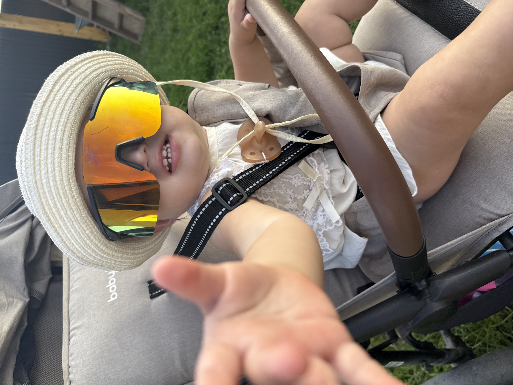 A baby is sitting in a stroller outdoors. The baby is wearing a straw hat and large, mirrored sunglasses that cover most of its face. The baby has a pacifier attached to its clothing. One of the baby's hands is reaching out towards the camera. The stroller is grey and has a brown handlebar. In the background, there is green grass and a wooden fence. The image appears to have been taken in the city of Podshivalovo, Russia.