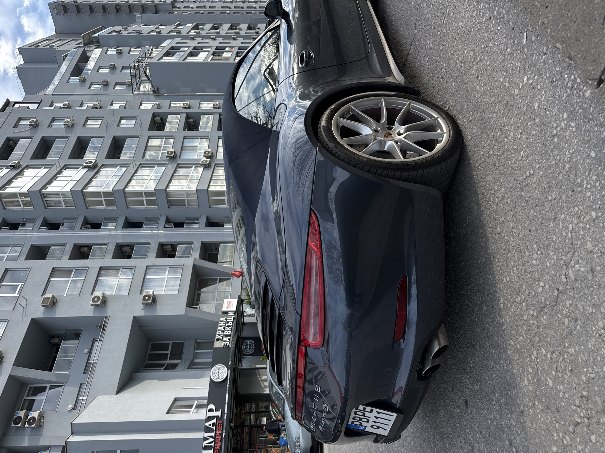 The image shows the rear of a dark-colored Porsche 911 Carrera S parked on a street in Plovdiv, Bulgaria. The license plate is visible and reads "PBPE 9111". Behind the car is a tall, modern building with numerous windows and air conditioning units. In front of the building is a storefront with signage in Cyrillic, including the word "MAPMARKET." A partial view of another vehicle is visible behind the Porsche. The street appears to be paved and in good condition. The sky is partly cloudy.