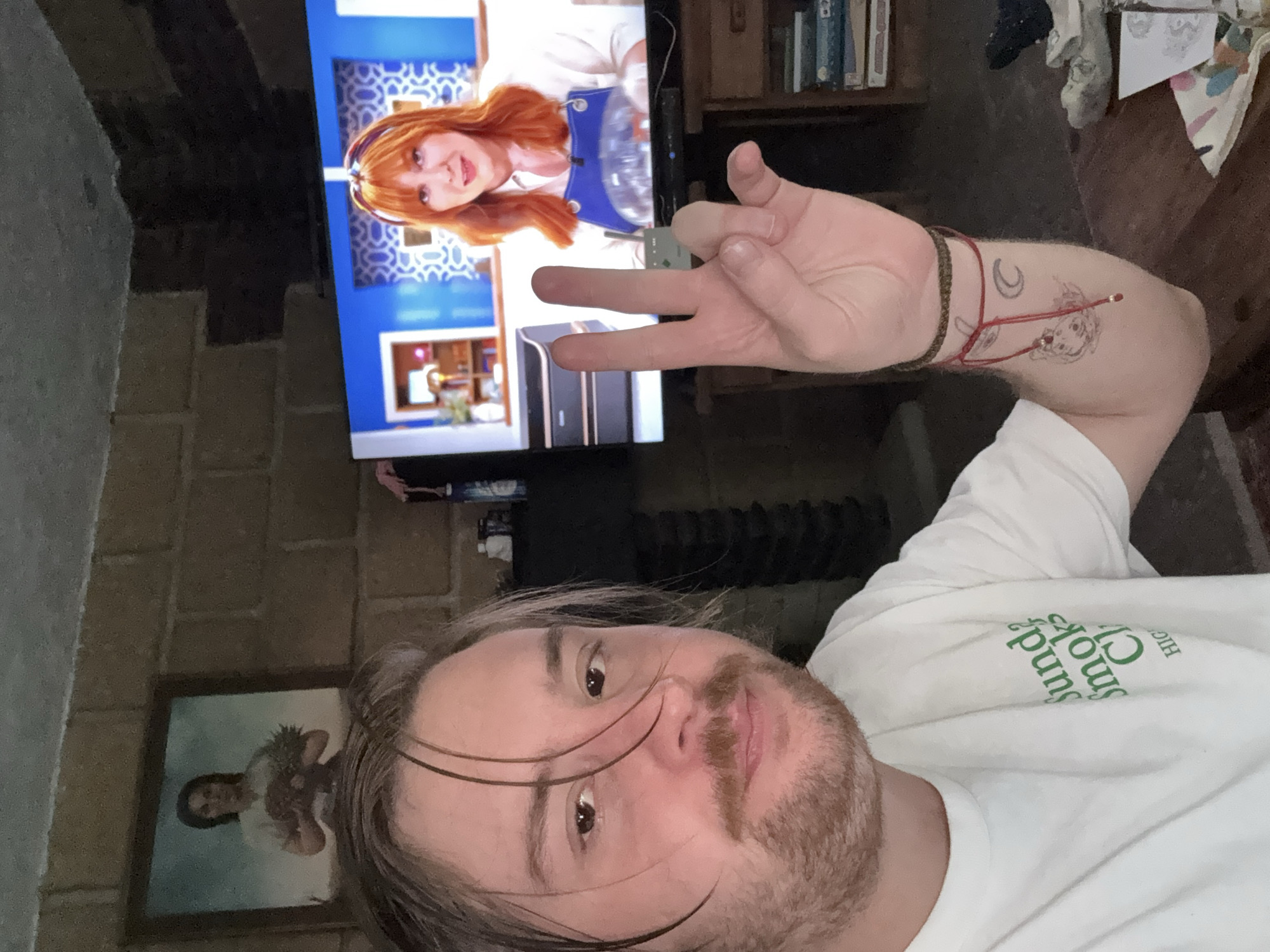 A man is taking a selfie indoors. He is lying down and is wearing a white t-shirt. His arm is raised and his hand is making a gesture with two fingers extended. He has a bracelet on his wrist and a tattoo on his arm. Behind him, a television screen shows a woman with red hair. There is also a framed picture on the wall and a shelf with books. The location appears to be indoors, potentially a living room. The location in Cañada de Madero, Mexico cannot be determined from this image.