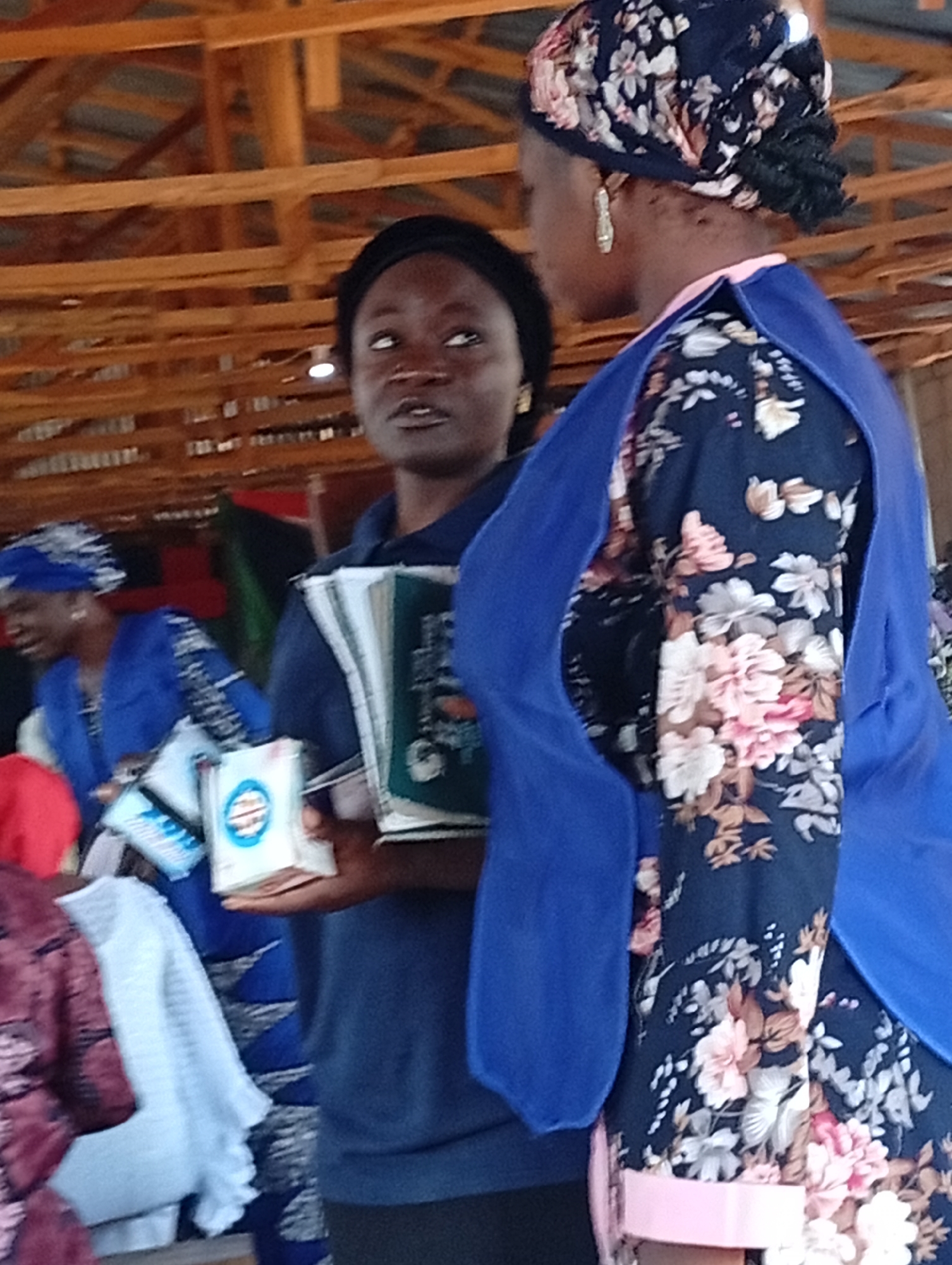 Two women are standing inside a wooden structure. The woman on the left is wearing a dark blue long-sleeved shirt and a black headscarf. She is holding a stack of books or pamphlets in her right hand. The woman on the right is wearing a dark blue floral print dress with a bright blue vest or apron over it. She has a patterned headscarf with pink, white, and black flowers. She is looking at the woman on the left. The structure appears to be a market or workshop with exposed wooden beams and roofing. The lighting is dim and natural.