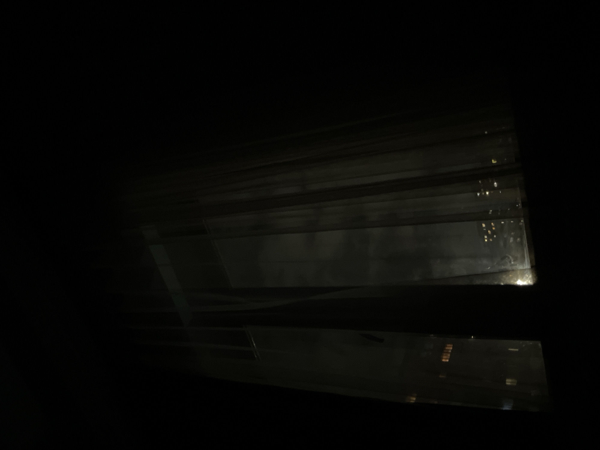The media file presents a very dark, nocturnal scene, dominated by deep shadows and minimal illumination. The primary visual elements are several dark, semi-transparent or reflective layers that occupy the foreground, arranged horizontally and slightly diagonally. These layers suggest a window structure, blinds, or louvers, creating an obscured and layered perspective.
Through and reflected upon these foreground elements, numerous small, scattered points of artificial light are visible in the background. These lights, some appearing as small rectangles, signify an urban environment at night, likely originating from distant buildings, streetlights, or other city illuminations. The scene captures an interior view looking out onto a dark cityscape.
No people or explicit activities are visible; the image is a static representation of a nocturnal landscape. The time of day is clearly night, emphasized by the pervasive darkness and the reliance on artificial light sources. No specific weather conditions can be determined. The overall impression is one of quietude and observation from an interior space into an illuminated urban expanse. There is no visible text in the image. Based on the provided context, the location depicted is Yerevan, Armenia.