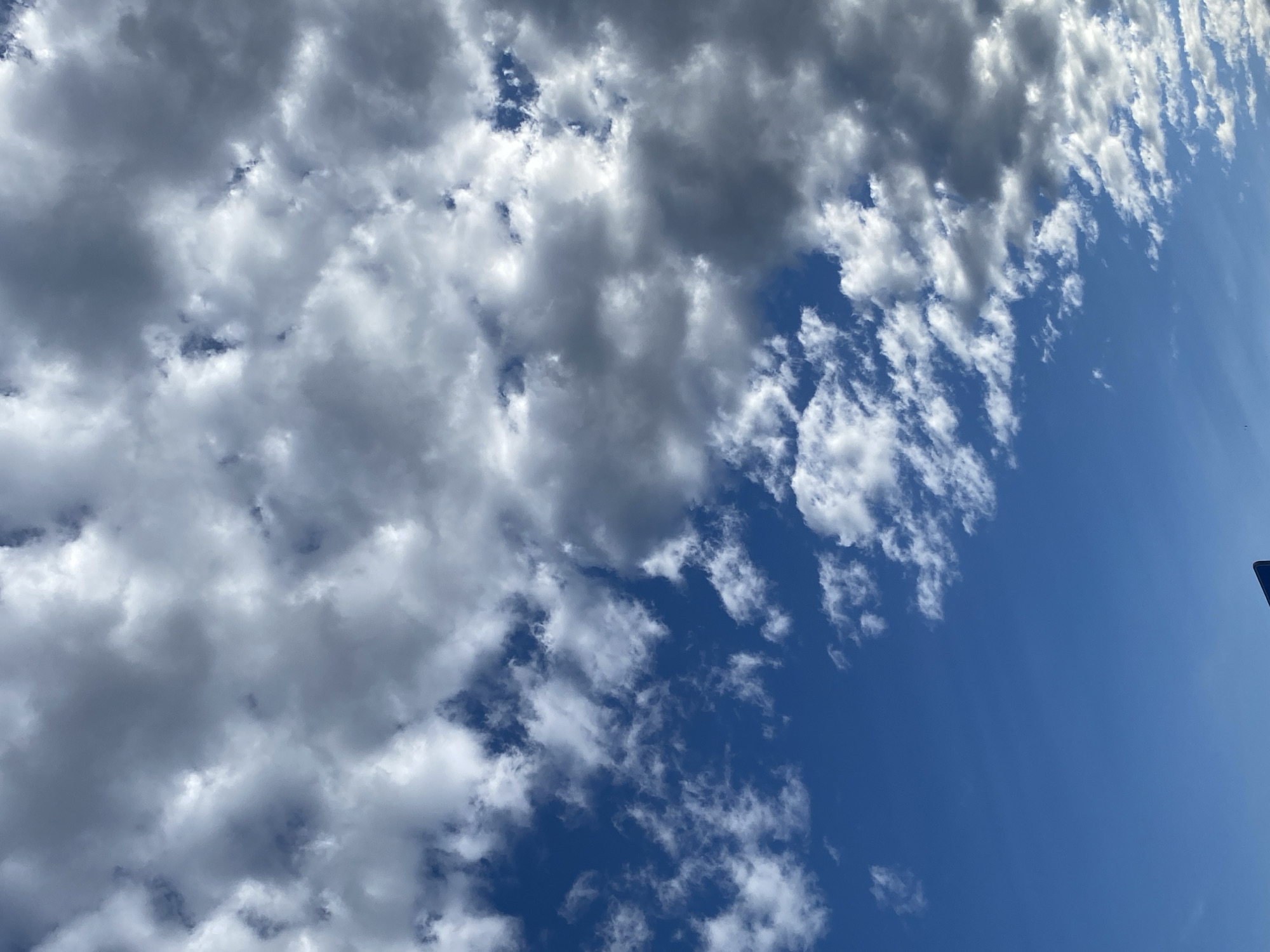 The image shows a bright blue sky filled with a mix of white and gray clouds. Some clouds are thin and wispy, scattered across the upper part of the frame, while others are thicker and more dense, appearing on the right side of the image. The lower portion of the sky is clear blue. A small portion of a dark blue sign with a white edge is visible at the very bottom of the image. The scene appears to be an outdoor view, likely from ground level, looking upwards. The context provided places this scene in the city of Dobieżyn, Poland.