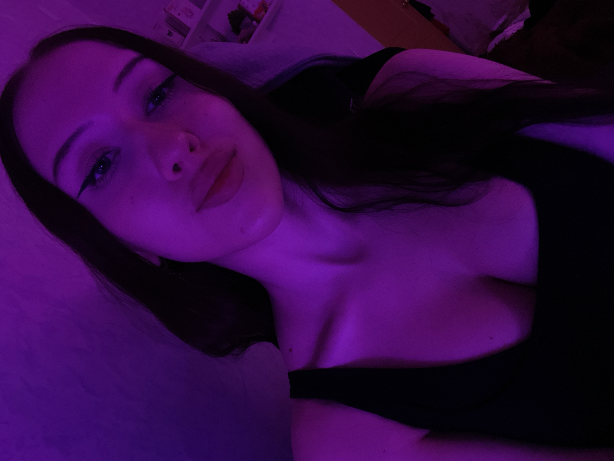 The image shows a young woman with long, dark hair, wearing a black top. She is illuminated by purple lighting, which casts a strong hue over the scene. The woman is looking towards the camera, with her head tilted slightly. Her makeup includes winged eyeliner. The background is indistinct due to the lighting conditions and focus. There is a shelf with some items visible on the right side of the image. The image does not contain information that allows for determination of the specific location in Votkinsk, Russia.