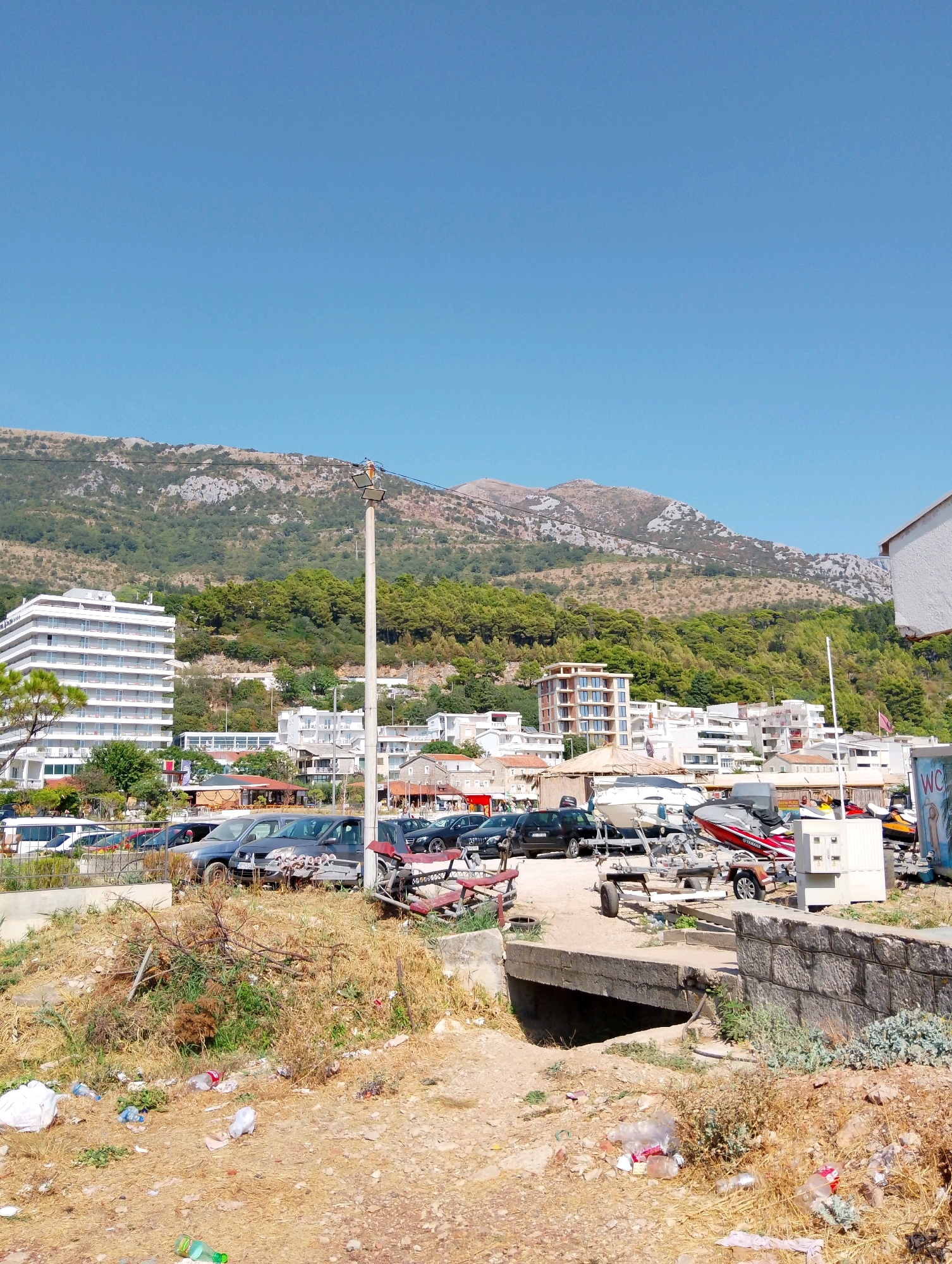 The image depicts a scene in Sutomore, Montenegro, characterized by a clear blue sky and a mountain range in the background. In the mid-ground, there is a parking area filled with various vehicles, including cars and some watercraft on trailers. Several buildings are visible, some modern and white, while others appear to be older. In the foreground, the ground is unpaved and littered with dry vegetation and trash, including plastic bottles and bags, suggesting an unmaintained area. A small concrete bridge or culvert crosses a dry ditch. A street light stands prominently on the right side of the frame. The overall impression is of a coastal town, possibly near a beach or marina, with a mix of modern development and a somewhat neglected foreground.