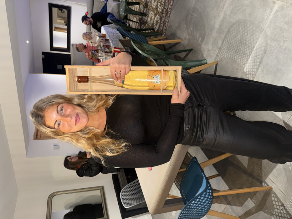 A fair-skinned woman with long blonde hair holds a wooden box containing a large bottle of white wine. She wears a black long-sleeved shirt and black pants. The photo is taken indoors. A table with chairs, other people, and interior décor are visible in the background.