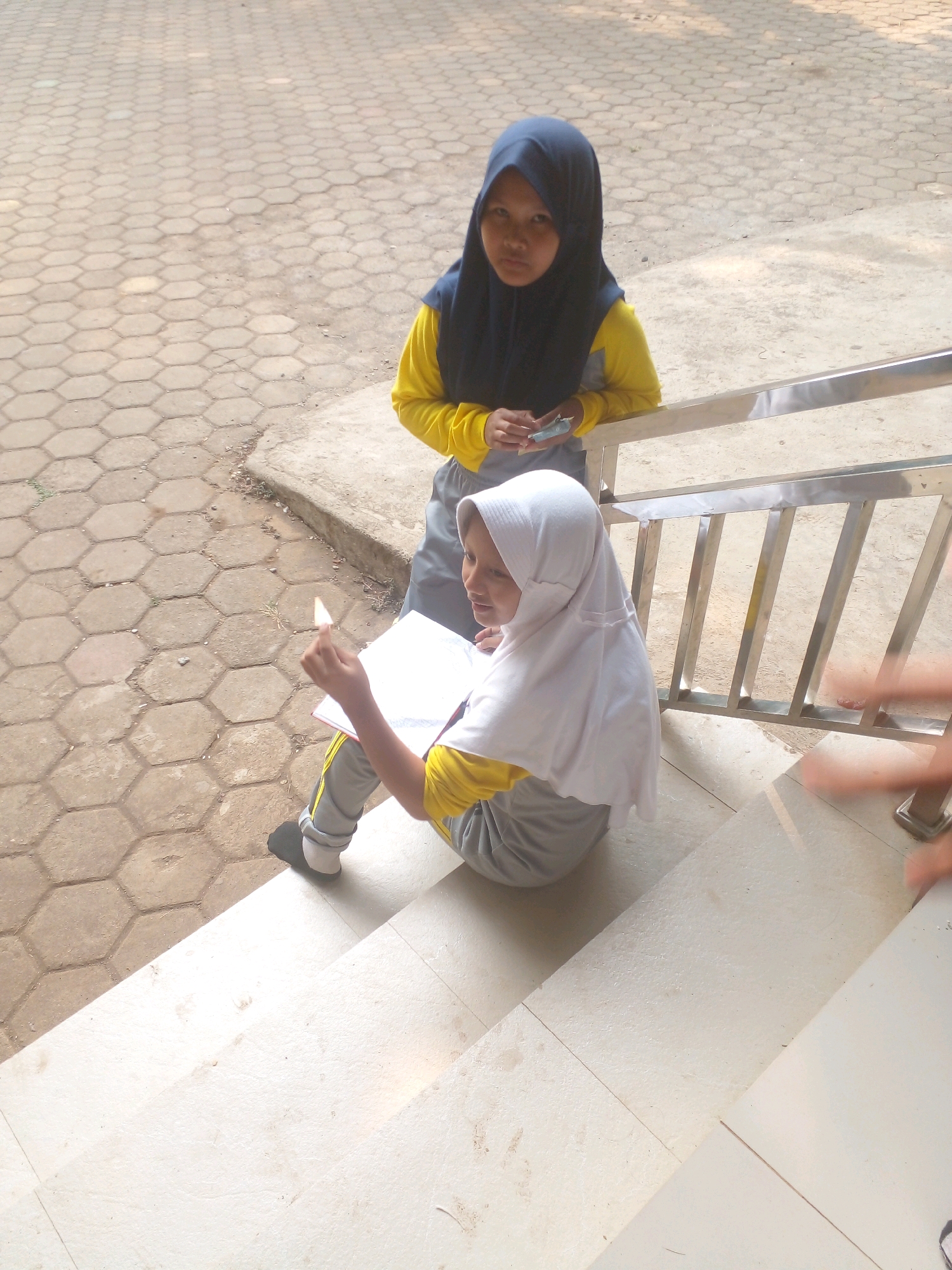 Two young females, likely students, are positioned on concrete steps outdoors. The female on the lower right, wearing a white hijab and a yellow long-sleeved shirt with gray pants, is seated and appears to be reading or writing in an open notebook. She holds a small white object, possibly a pencil or pen, in her right hand. The second female, standing on a higher step to the left, wears a dark blue hijab and a yellow long-sleeved shirt with gray pants. She is looking towards the viewer and holding a small rectangular object, possibly a phone or a small book, in her hands. The environment suggests a public outdoor space, possibly a school campus or a public building in Banyuputih, Indonesia, given the distinct hexagonal patterned paving stones visible in the background and the presence of a metal railing. The lighting indicates it is daytime, with shadows cast on the ground.