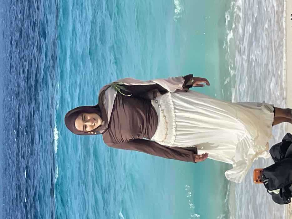 A female subject stands on a sandy beach in Zanzibar, Tanzania, facing forward with a slight smile. She wears a dark brown long-sleeved top, a long off-white pleated skirt, a brown hijab, and a light-colored sheer fabric draped over her left shoulder. Gold-colored drop earrings and a pearl-beaded chain belt are visible accessories. A small green plant or leaf is tucked into the sheer fabric on her left shoulder. Her lower legs are covered, possibly by leggings, beneath the skirt, which appears wet at the hem from the ocean water. In the lower-left foreground, a black bag supports an orange-cased smartphone, which is angled towards the subject, indicating it is recording or photographing. The background features the ocean with distinct bands of turquoise and deeper blue water, showing visible waves and ripples under clear daylight conditions.