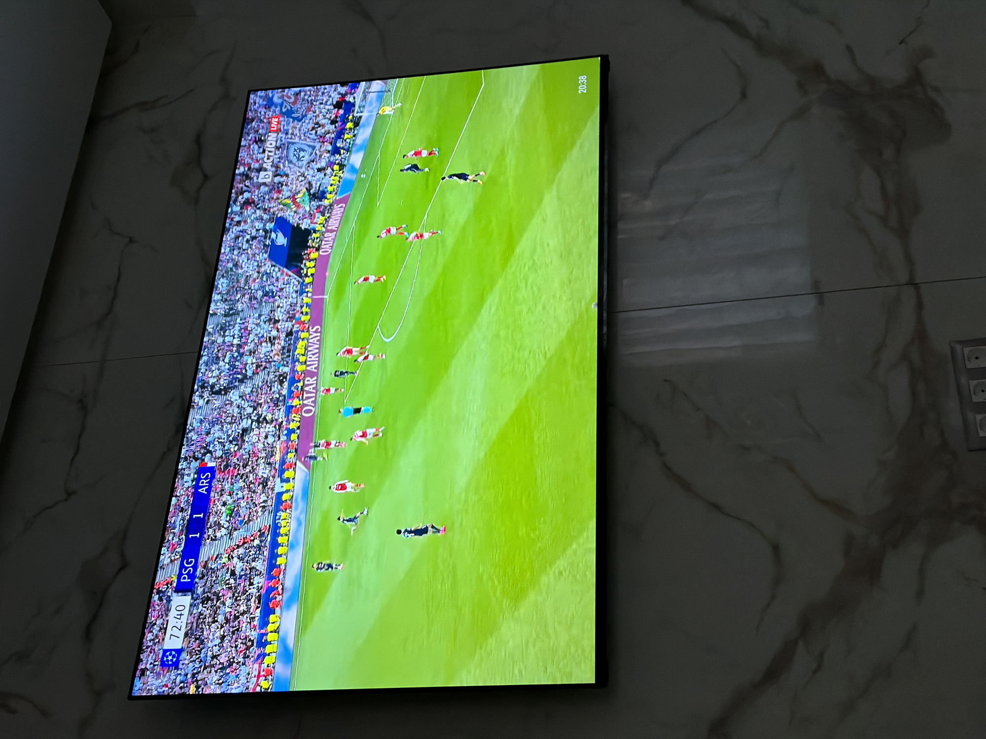 The image shows a television displaying a soccer game between Paris Saint-Germain (PSG) and Arsenal (ARS). The score is tied 1-1 at 72:40. The game appears to be live. The TV is mounted on a marble-patterned wall. The location is likely a residential setting in Asenovgrad, Bulgaria, as that information was provided. A light-colored wall is visible to the left of the TV, and an electrical outlet is on the wall to the right.