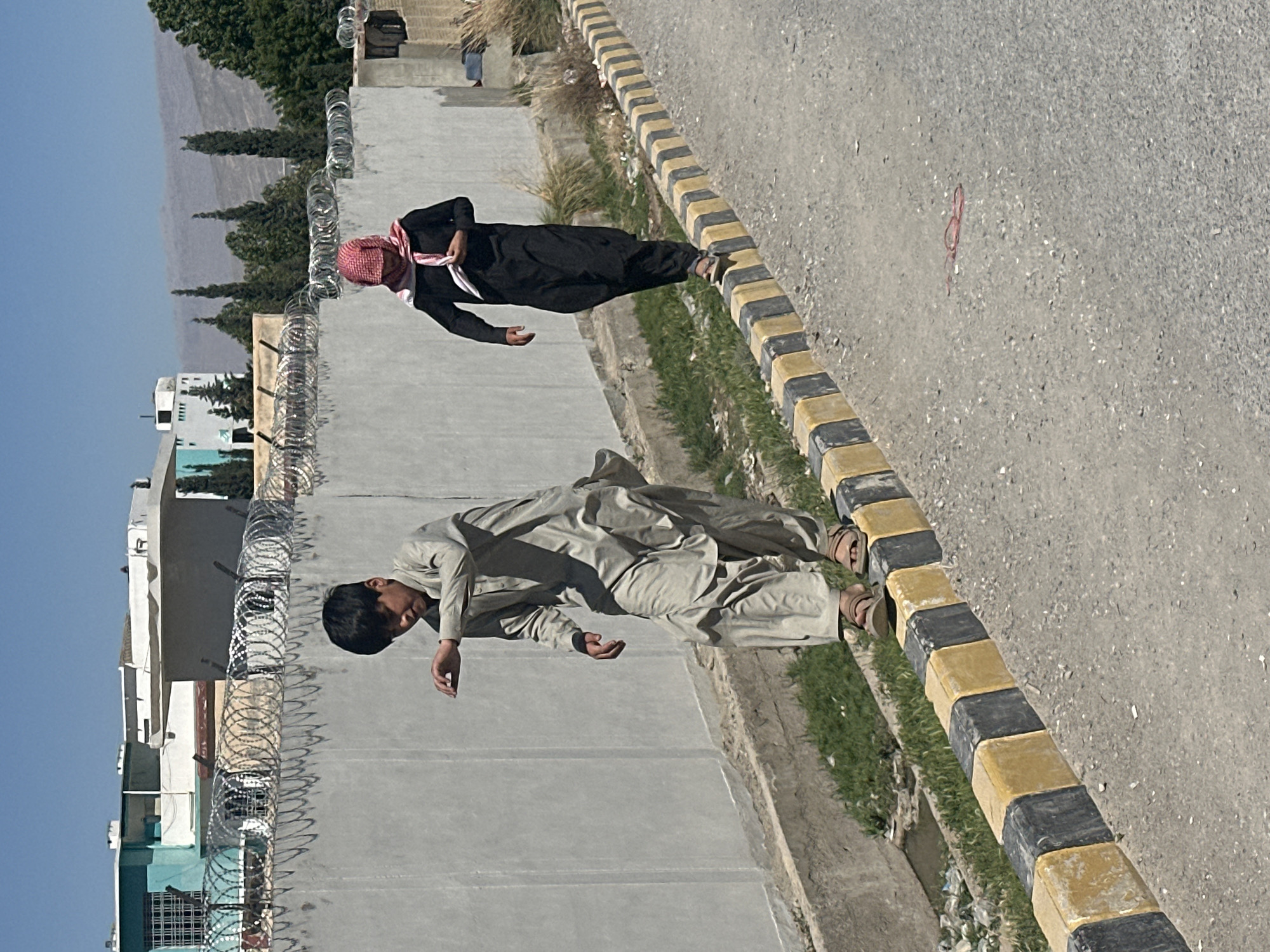 Two men stand outside a gated compound with barbed wire in Quetta City Tehsil, Pakistan. One man wears a brown shalwar kameez, while the other wears a black robe and a red and white head covering. They stand near a road separated by a curb with alternating yellow and black markings. Buildings and distant mountains are visible in the background.