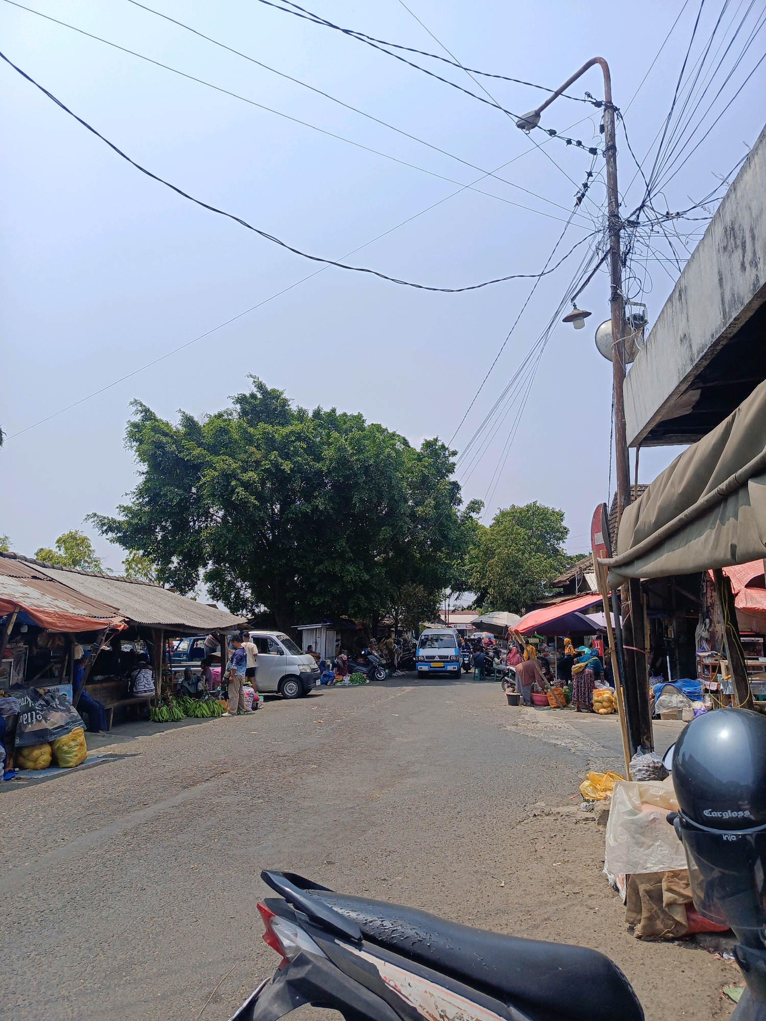 This is a medium-angle, eye-level, outdoor photograph of a street market in Sukowinangun, Indonesia. The scene is captured on a clear, bright day with a pale blue sky and a few scattered power lines crisscrossing overhead. The foreground features the back of a black motorcycle with a grey helmet placed on its seat.
The street is paved with asphalt and appears to be a two-lane road with vendors set up on both sides. On the left side of the street, there are several stalls with corrugated metal roofs, some covered with tarps. Various goods are displayed, including large sacks of produce, possibly bananas, and other items wrapped in plastic. Several people are seen interacting at these stalls.
On the right side of the street, a more established building with a concrete facade and a tarpaulin awning lines the road. More vendors are present here, some under umbrellas, selling a variety of items that appear to be food or household goods. People are actively browsing and making purchases.
In the middle ground, a silver minivan is parked on the street, and a blue van is driving away. A large, mature tree with dense green foliage stands prominently behind the stalls on the left, providing shade. The overall environment suggests a bustling local market, likely a regular occurrence in this Indonesian town. The image captures the daily life and commerce of the community.