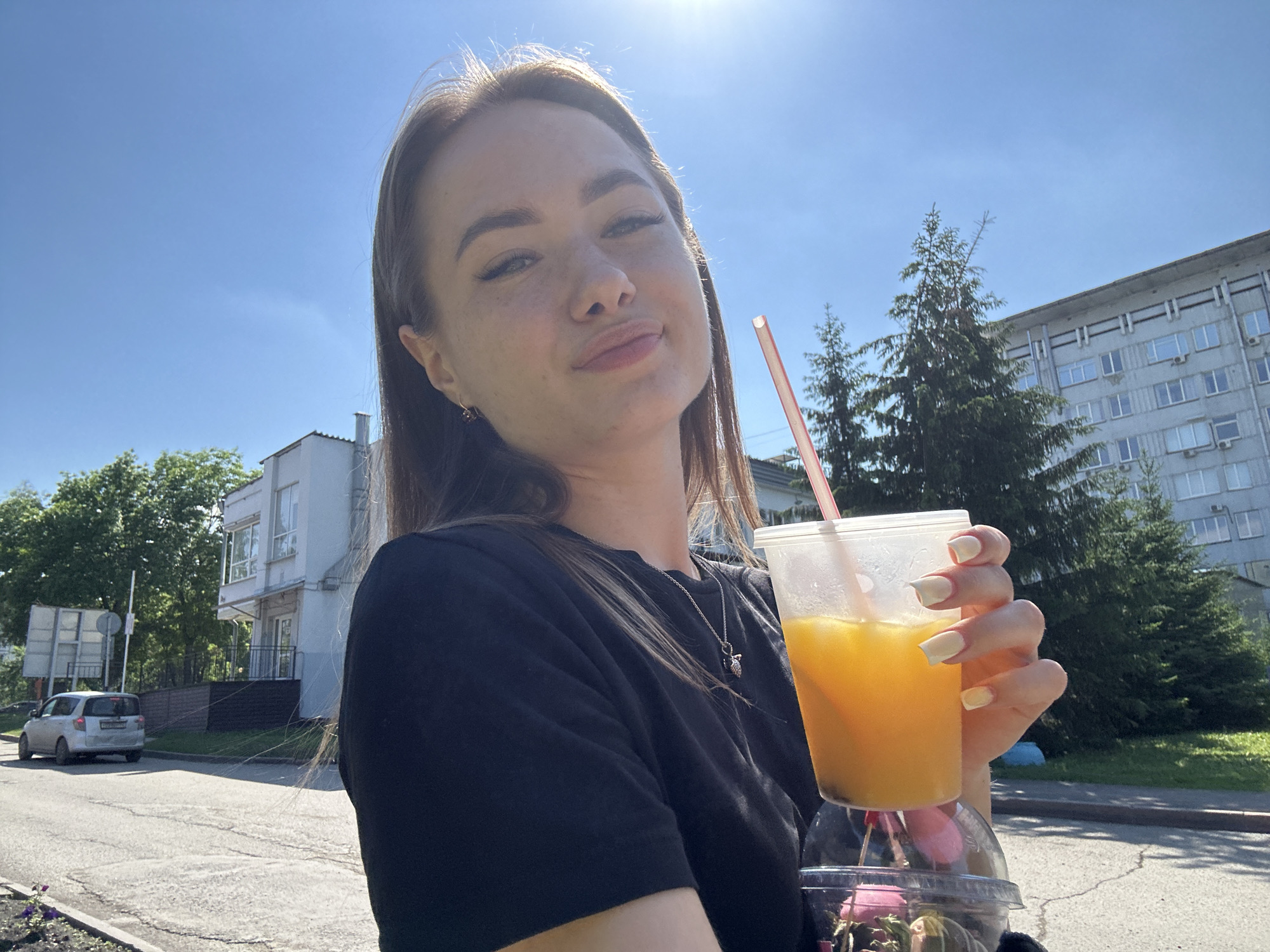 A young woman with long brown hair, wearing a black t-shirt and a silver necklace, smiles at the camera while holding a clear plastic cup filled with orange juice and ice. She has long, light-colored fingernails. The background shows a blue sky with the sun shining brightly, some trees, and buildings. A white car is parked on the street to the left of the frame.
