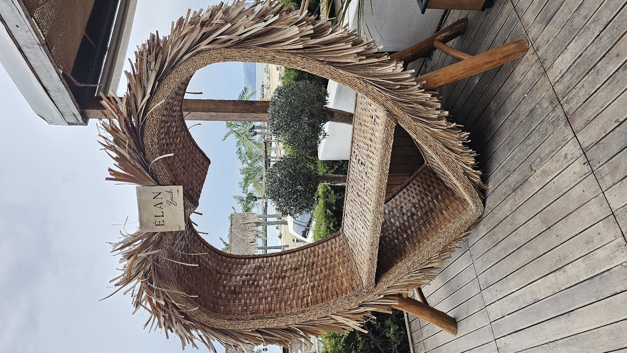 The image depicts a scene at "ÉLAN Beach," as indicated by a sign in the foreground. The location is in Varna, Bulgaria, given the context. The scene includes a unique, woven structure resembling a chair or shelter, made of natural materials like straw or reeds. It has a curved, almost heart-like shape with a thatched fringe. The structure sits on a wooden deck, which is oriented horizontally in the image. In the background, palm trees, manicured shrubs, and glimpses of the sea are visible through the opening of the woven structure. A white building is partially visible behind the foliage. The sky is overcast.