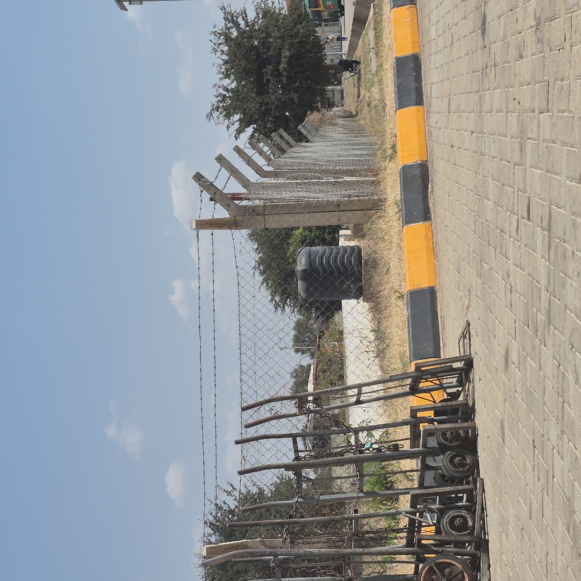 In Dodoma, Tanzania, a scene unfolds with a focus on utility and outdoor elements. The foreground features a paved area with interlocking brick pavers, suggesting an outdoor workspace or thoroughfare. To the left, a collection of industrial trolleys, characterized by their metal frames and wheels, are stacked and leaning. Chains are visible, some attached to the trolleys and others hanging, hinting at their use for moving heavy loads.
Beyond the trolleys, a fence constructed of chain-link mesh and topped with barbed wire marks a boundary. Concrete posts support the fence, with some showing signs of wear. Behind the fence, a large, black, ribbed water tank stands on a concrete base. Further back, sparse, dry vegetation and a few trees create a natural backdrop. In the distance, a green and yellow bus is partially visible, indicating ongoing activity or transportation. The sky above is a clear, light blue with a few scattered white clouds. The overall impression is one of an organized, possibly industrial or logistical, outdoor area in a dry climate.
