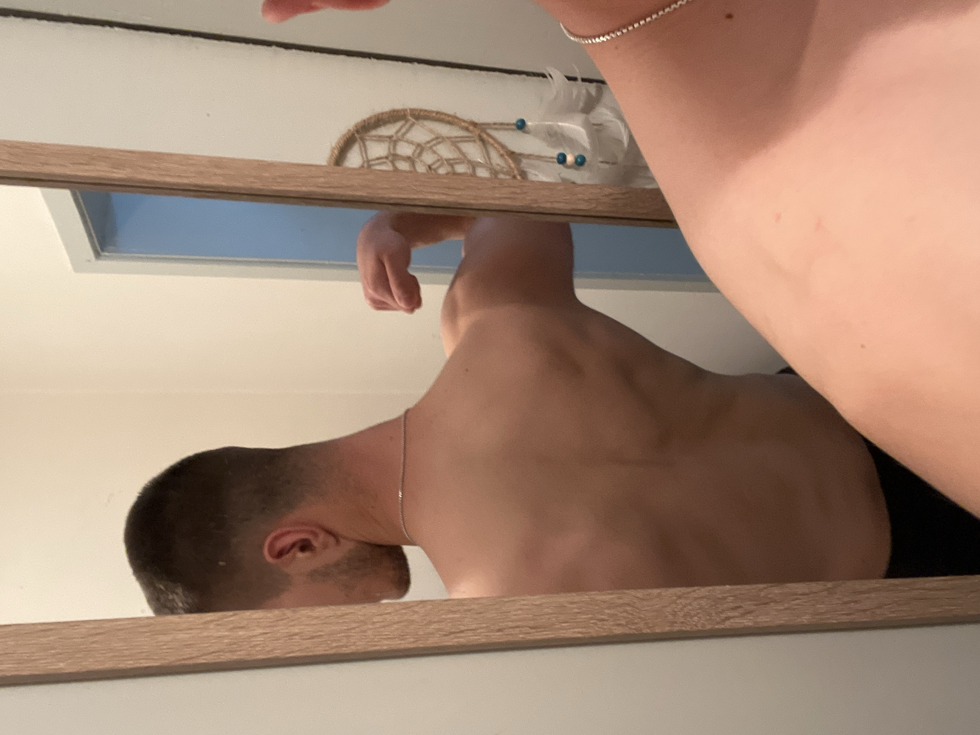 A mirror reflects a man's shaved head and bare back, wearing a thin chain necklace, as he is shirtless. A woman's upper arm with a silver bracelet is partially visible in the upper right corner. In the background, above the mirror, there is a dreamcatcher. The setting is an indoor space with a window visible in the reflection. The location is Unknown, Unknown.