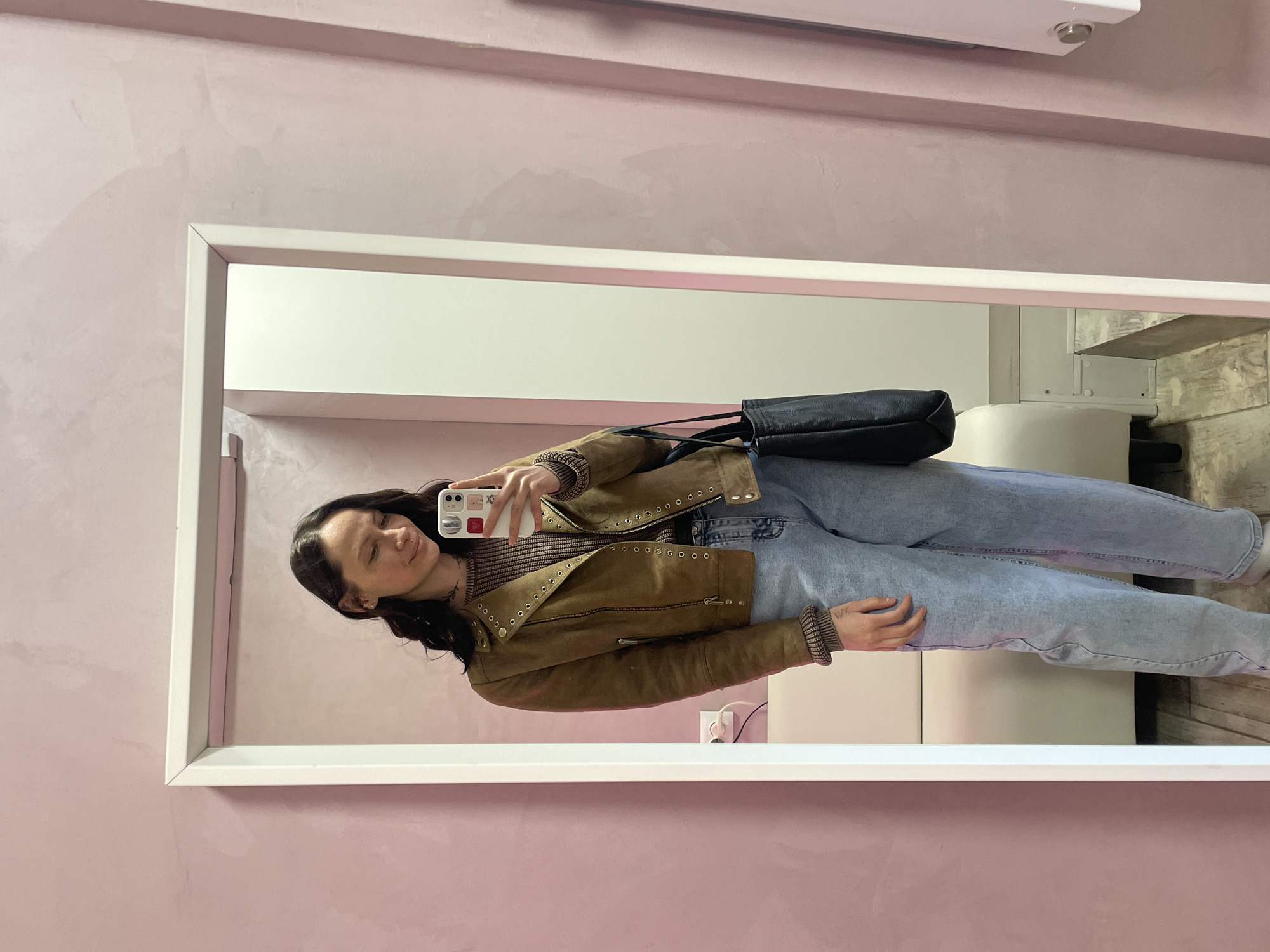 A woman stands in front of a mirror, taking a selfie. She wears a brown jacket, a brown and beige striped top, and light blue jeans. A black purse hangs over her shoulder. The mirror has a white frame and is set against a pink wall. The reflection shows a white wall and a cushioned seat. The location is Izhevsk, Russia.