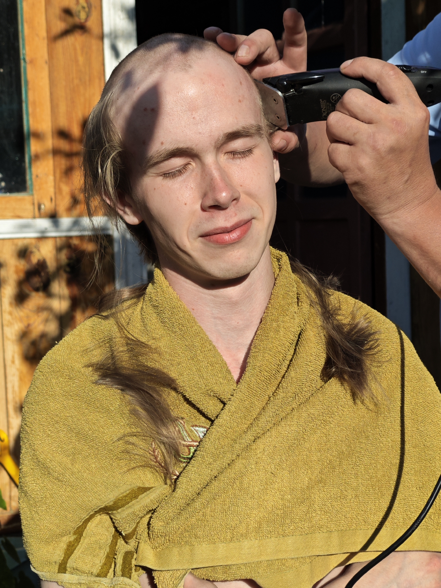 A young adult with closed eyes and a faint smile is having their head shaved with an electric razor outdoors. They are wearing a yellow terrycloth robe and have longer hair on the sides and back of their head. Sunlight casts shadows on their face and head. The background shows a wooden door or wall and foliage, suggesting a residential setting, possibly in the city of Fokino, Russia.