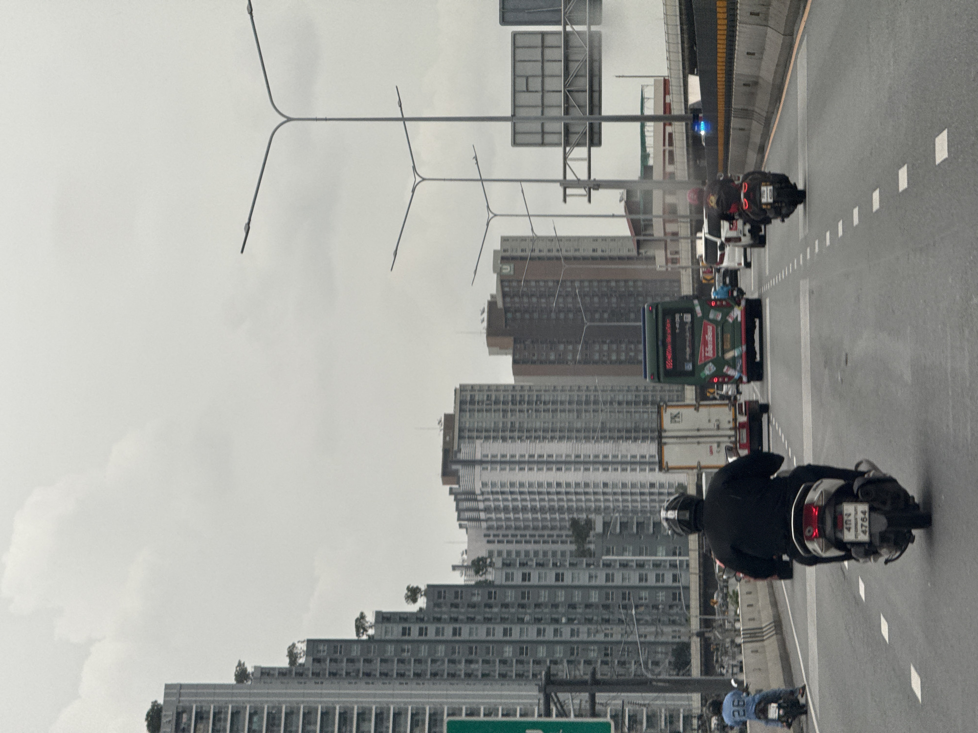 In Bangkok, Thailand, on a highway on an overcast day, a person wearing a black helmet and black clothing rides a white scooter. The scooter has a license plate with Thai script and the number "4764". The scooter is on the right side of the road, in the middle lane. In front of the scooter, there is a white truck and a green bus. To the left, a person wearing a blue shirt with the number "26" on it rides a black scooter. In the background, there are tall buildings. The sky is cloudy.