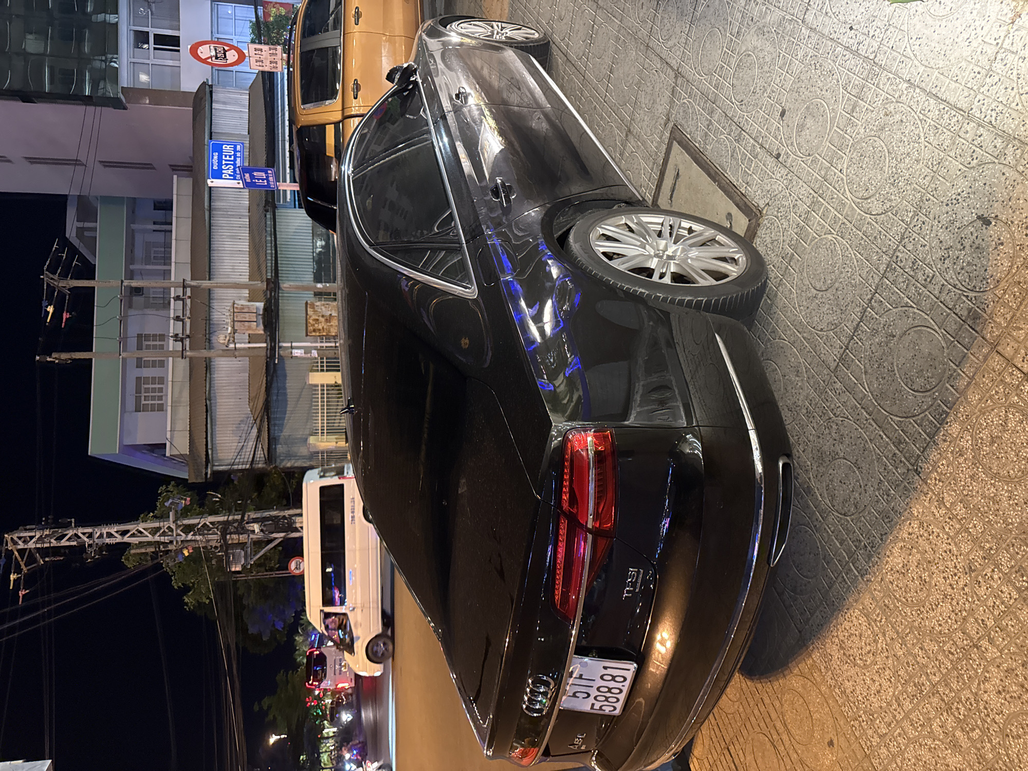 The image captures a nighttime scene on a city street in Nha Trang Ward, Vietnam. The primary subjects are two vehicles: a sleek black Audi sedan and a larger, light-colored van. The Audi is parked on a tiled sidewalk, its polished black surface reflecting the surrounding lights. Its rear taillight assembly is prominent, and a license plate with the characters "51F 5888.81" is clearly visible. The van is parked further down the street, partially obscuring a traffic light and a dense network of electrical wires overhead.
The setting appears to be urban, with buildings visible in the background. One building has a sign that reads "DUONG PASTEUR" and another "LE LOI," indicating street names. There are also traffic signs, including one with a circular red symbol and some text that is difficult to decipher due to the image quality and rotation. The time of day is clearly night, as evidenced by the dark sky and artificial lighting. The weather seems calm, with no indication of rain or wind. The overall mood is that of a typical urban street scene at night, with the vehicles suggesting ongoing activity, possibly related to transportation or commerce. The reflection on the Audi suggests the pavement is wet, but this is not definitively confirmed by other visual cues.