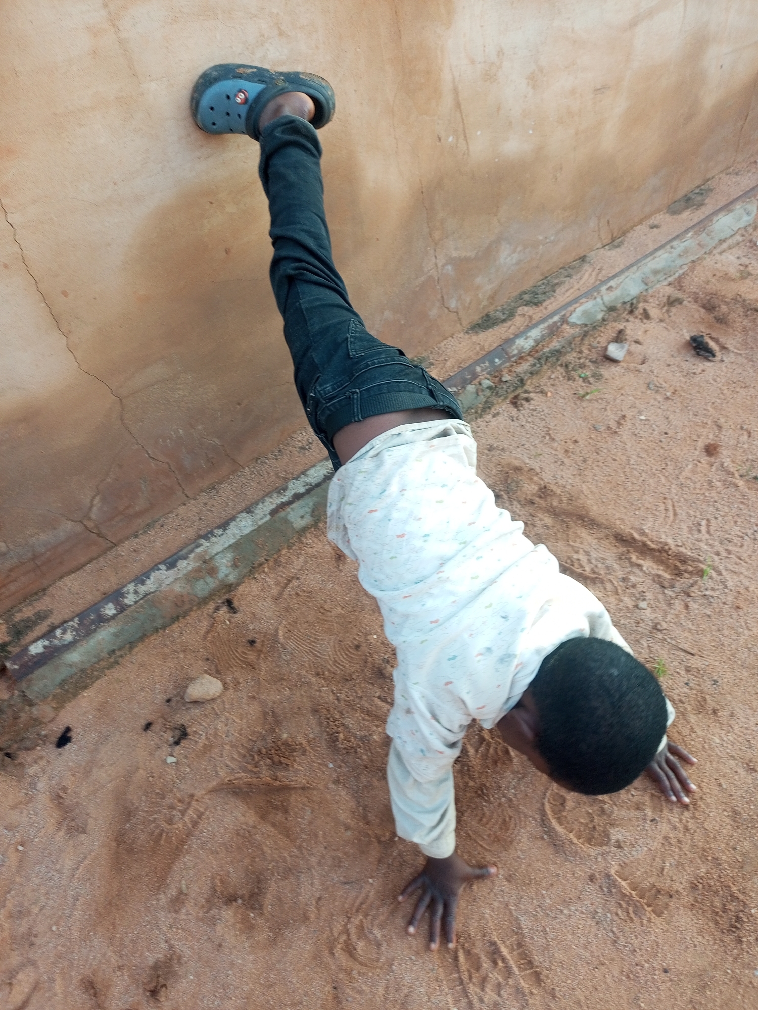 A young Black child, wearing a light-colored shirt with a subtle pattern and dark jeans, is positioned in a handstand against a textured, light brown wall. The child's right foot, clad in a dark blue Croc shoe, is pressed firmly against the wall. The child's body extends downwards from the wall, with hands on the sandy ground. The environment appears to be an outdoor setting with a sandy floor, possibly in a courtyard or along a pathway, near what looks like a concrete foundation or drainage channel. The scene is set in Tempure, Nigeria.