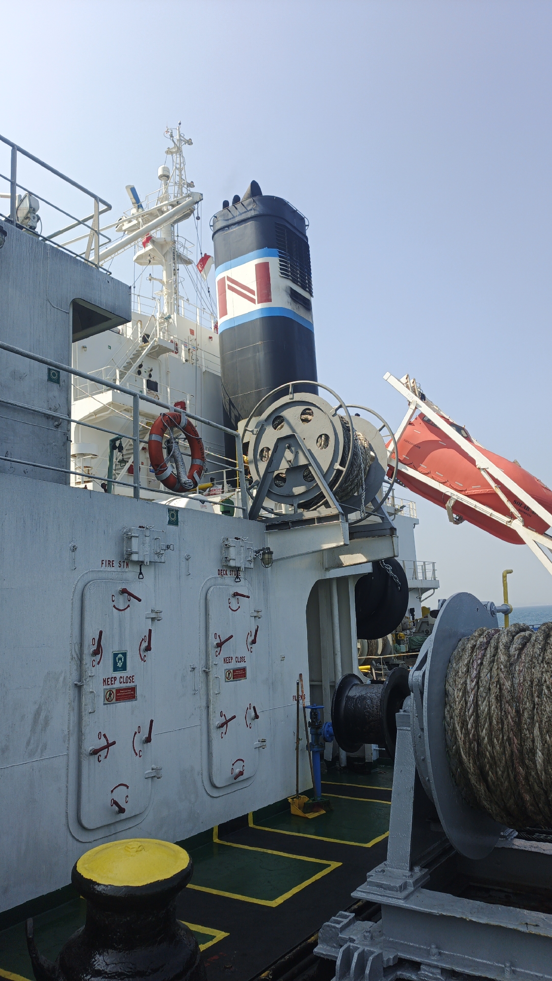 This is a photograph taken outdoors on the deck of a ship, likely in or near Singapore. The sky is clear and blue. On the left side of the frame, a large white structure, part of the ship's superstructure, is visible. Below this, on the hull, are several white doors with red handles and cautionary signs like "KEEP CLOSE". A life preserver ring is hanging on a railing above these doors. Dominating the center of the image is a tall, black smokestack with red, white, and blue markings, including what appears to be a red letter "N" on a white stripe. To the right of the smokestack, a red, enclosed lifeboat is mounted on a davit. In the foreground, a large winch with thick rope wound around it is prominently displayed, painted gray. The deck itself is dark green with yellow safety markings. The overall impression is one of a working vessel, possibly docked or at sea.