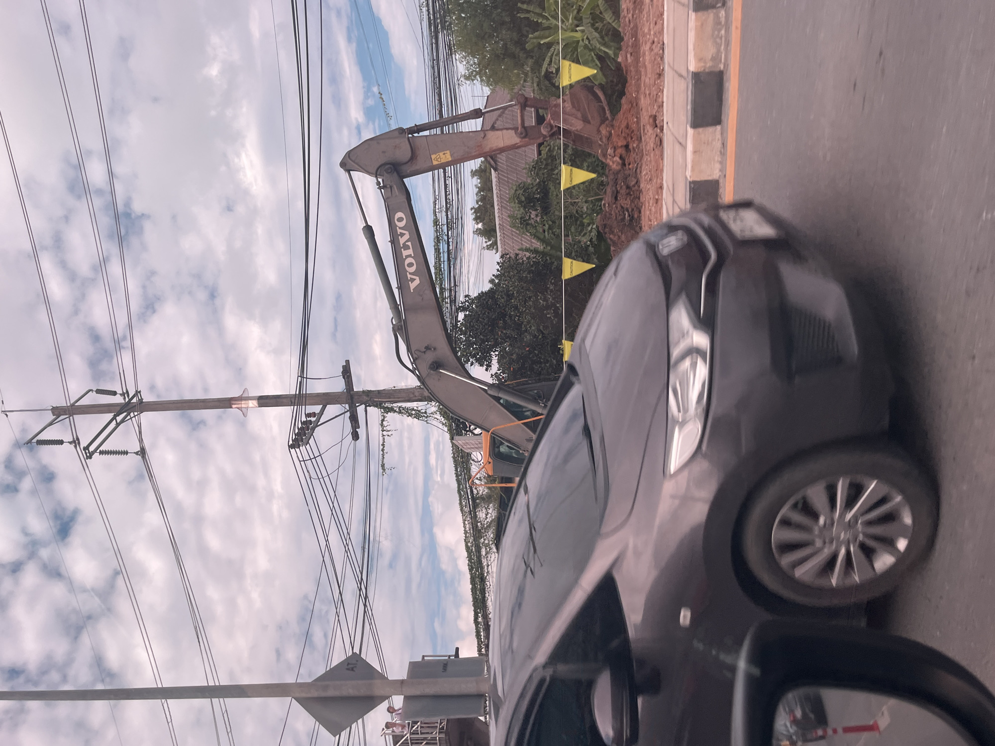In Rawai, Thailand, a dark grey car drives past a construction site. The car is in motion and partially obscures the view of the site. In the background, a large grey Volvo excavator is visible, with its arm extended over a pile of dirt. Yellow "CAUTION" flags are placed along the edge of the construction area. Overhead, a dense network of electrical wires and utility poles crisscrosses the sky, under a cloudy sky with patches of blue. A street sign is visible on a pole to the left, and two individuals can be seen standing on an elevated platform behind it. The foreground shows the car's hood and windshield, with the driver's side mirror reflecting a portion of the road and a red traffic cone.