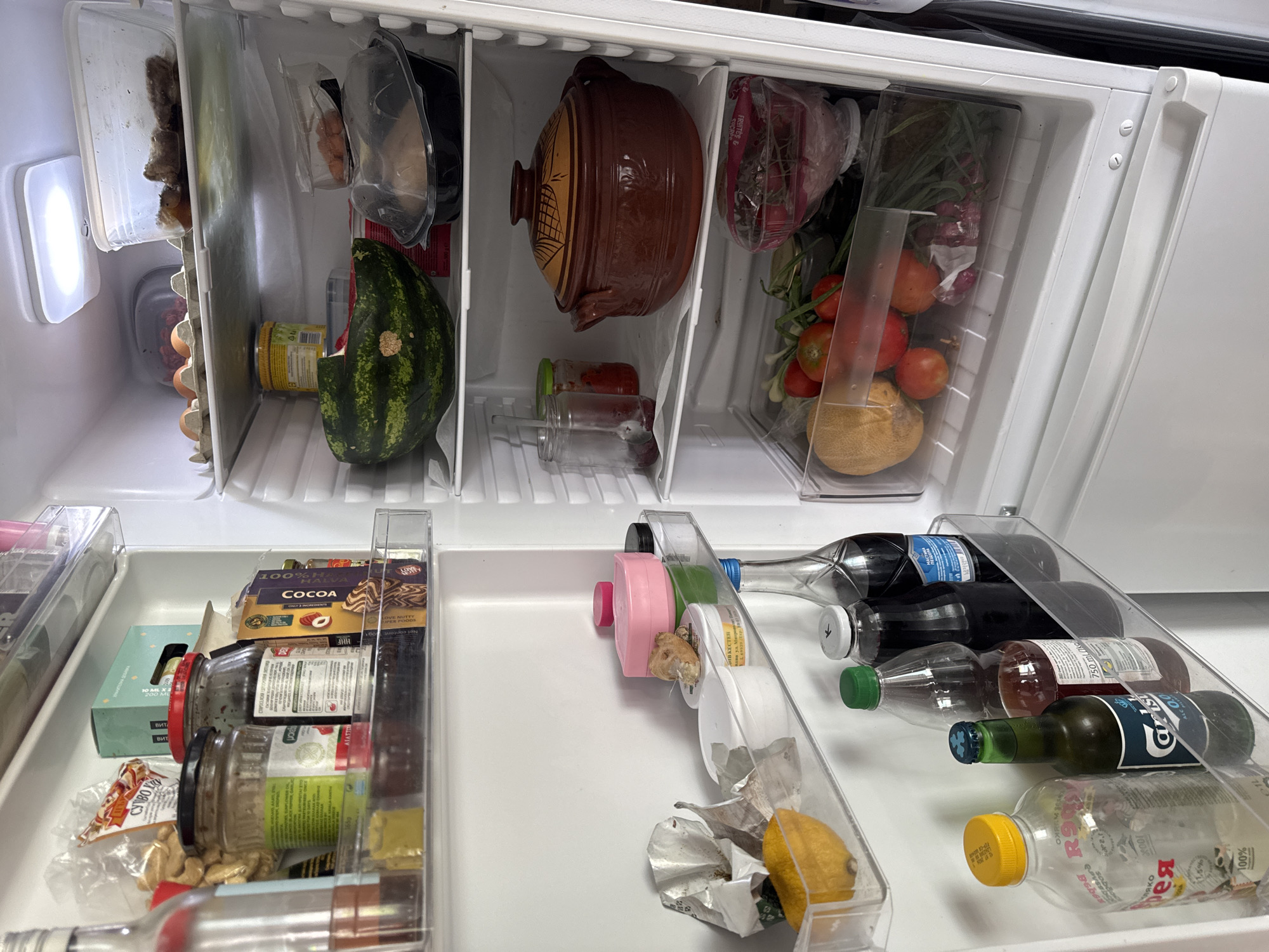 The image shows the interior of a refrigerator. On the top shelf, there are eggs in a carton and a plastic container with food. The second shelf contains a jar of yellow food, a watermelon, and a brown clay pot with a lid. The third shelf has several jars and bottles of drinks, including a bottle of Carlsberg. The crisper drawer at the bottom contains a melon, tomatoes, and green onions. The refrigerator is located in a residential kitchen in Plovdiv, Bulgaria.