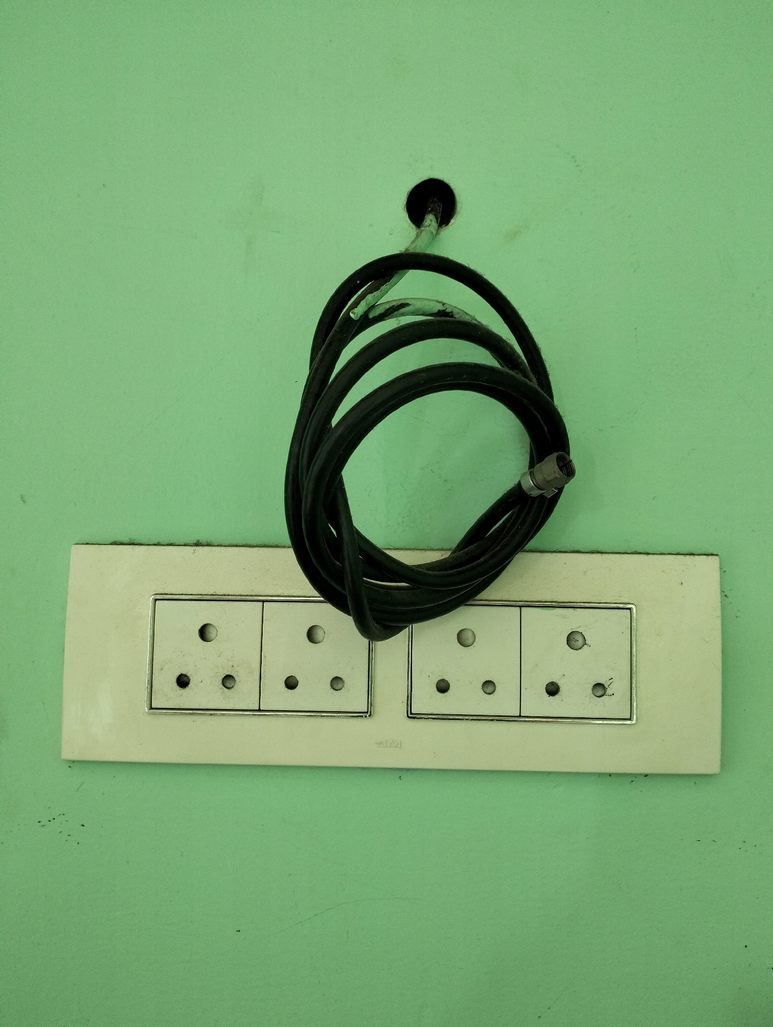A coiled black cable with a silver connector hangs from a hole in a light green wall. Below the cable, a white electrical outlet strip with two sets of three sockets is mounted horizontally on the wall. The outlet strip appears to be made of plastic and has a silver trim around the sockets. The background is a plain, slightly textured light green wall. No people are visible in the image. This scene is located in Sriperumbudur, India.