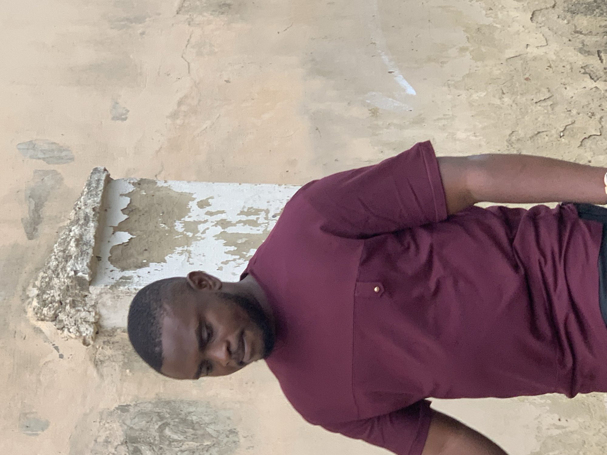 A man with a dark complexion and short hair is captured from the chest up, looking downwards and slightly to his left. He is wearing a maroon, short-sleeved shirt with a mock neckline and a horizontal seam across the chest, accented by a small, golden button on the left side. The background is a distressed, textured wall with patches of peeling paint and a weathered concrete structure to the left of the man. The scene appears to be outdoors, possibly in an urban environment like Abuja, Nigeria, given the architectural style of the background elements.