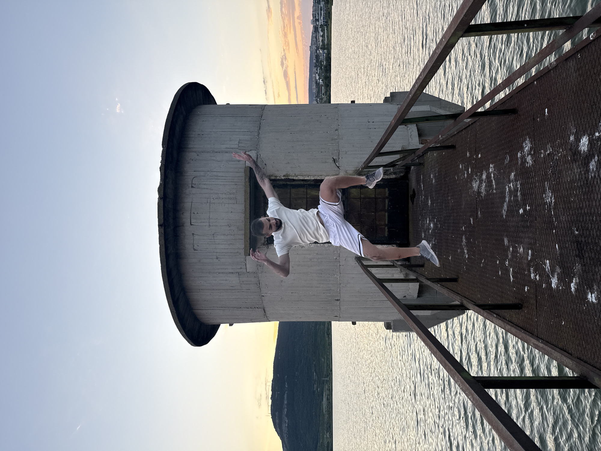 A man is captured mid-air, jumping on a rusty metal walkway leading to a cylindrical concrete structure situated on a body of water. The man, wearing a white t-shirt and white shorts with black stripes, is in the foreground, his body positioned horizontally as he leaps. The concrete structure, which appears to be a water tower, is centered behind him. The walkway extends from the viewer towards the structure. The background features a serene lake reflecting the soft hues of a twilight sky, with a distant hill and a city skyline visible on the horizon. The location is identified as the water tower on the Shumen plateau, near the city of Shumen, Bulgaria. The overall atmosphere is one of dynamic movement against a tranquil, natural backdrop.