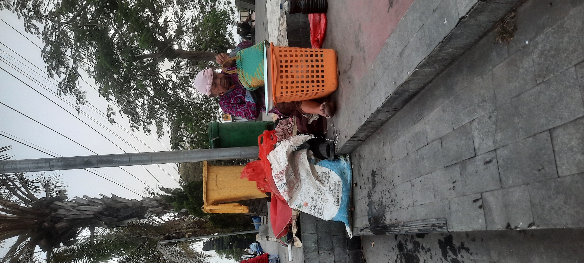 The image depicts an outdoor scene in the city of Kanigoro, Indonesia, likely along a sidewalk or public walkway. In the foreground, a person wearing a pink head covering and a patterned shirt sits on the ground next to a large white bag with blue lettering and an orange plastic basket. The person is holding a green woven bag. To the left of the person are two large plastic trash bins, one yellow and one green, with some red plastic items and a white bag on the ground nearby. The background features a tall palm tree, other trees with foliage, overhead power lines, and utility poles. Further in the background, several individuals are visible sitting or standing near a body of water. The ground is paved with gray stone tiles, with some areas appearing wet or stained. The overall atmosphere suggests a public area where people gather or pass through.