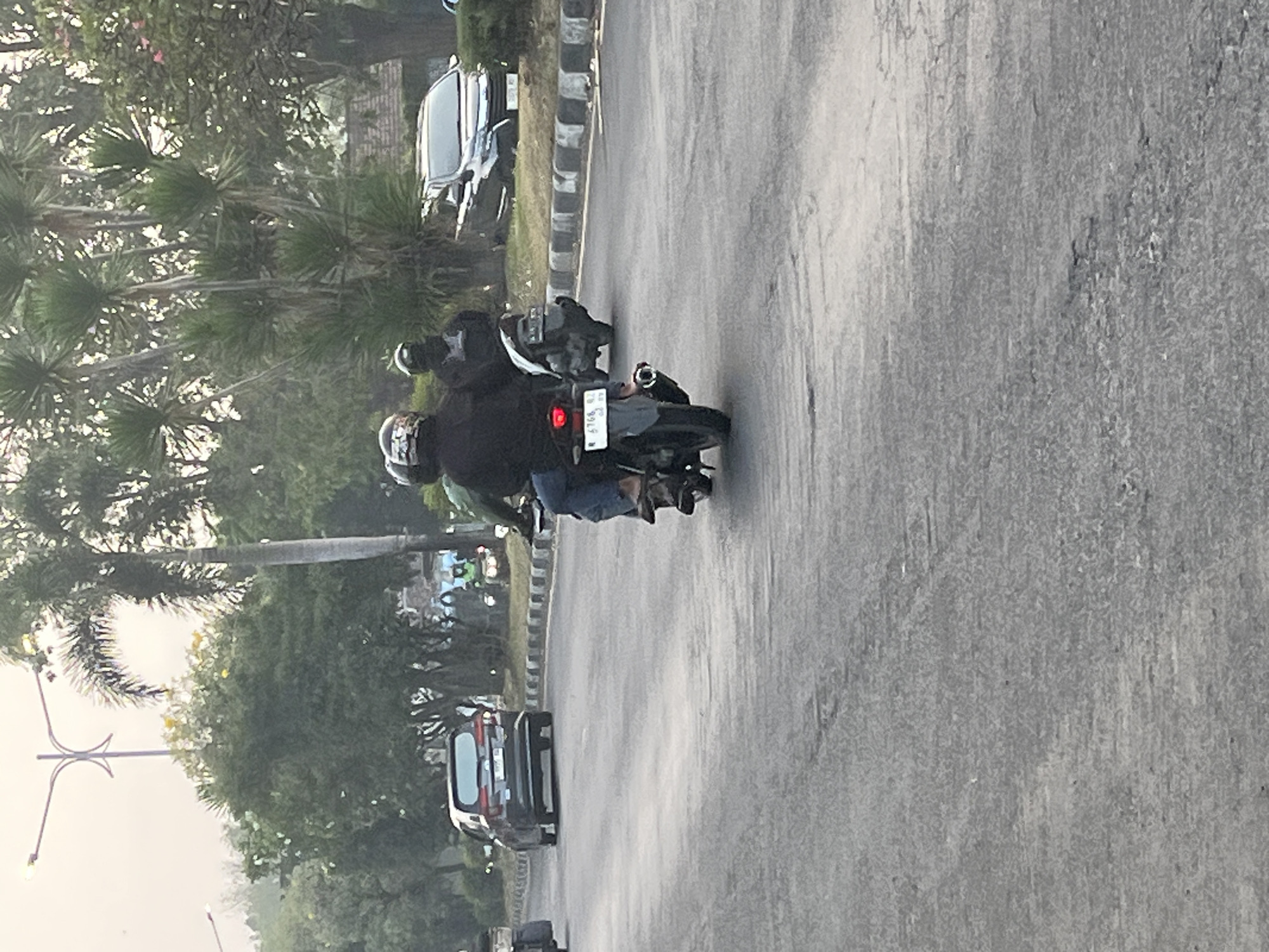 A medium shot captures a view of a street scene in Kludan, Indonesia, likely during daytime. The perspective is from behind a moving motorcycle, looking forward. The asphalt road is grey and textured, with some cracks visible. On the right side of the road, a sidewalk with black and white striped borders is present. Beyond the sidewalk, a line of palm trees and other lush green foliage forms a dense background.
In the foreground, the back of a motorcycle is visible, with its license plate reading "R 6768 QZ". The rider is wearing a black jacket and blue jeans, and the passenger is wearing a black helmet. Further ahead, another motorcycle is seen with two riders, both wearing helmets. To the left of the road, a dark grey car is driving away from the viewer. In the background, more trees and a glimpse of buildings suggest an urban or semi-urban environment. The sky is overcast, casting a diffused light over the scene.