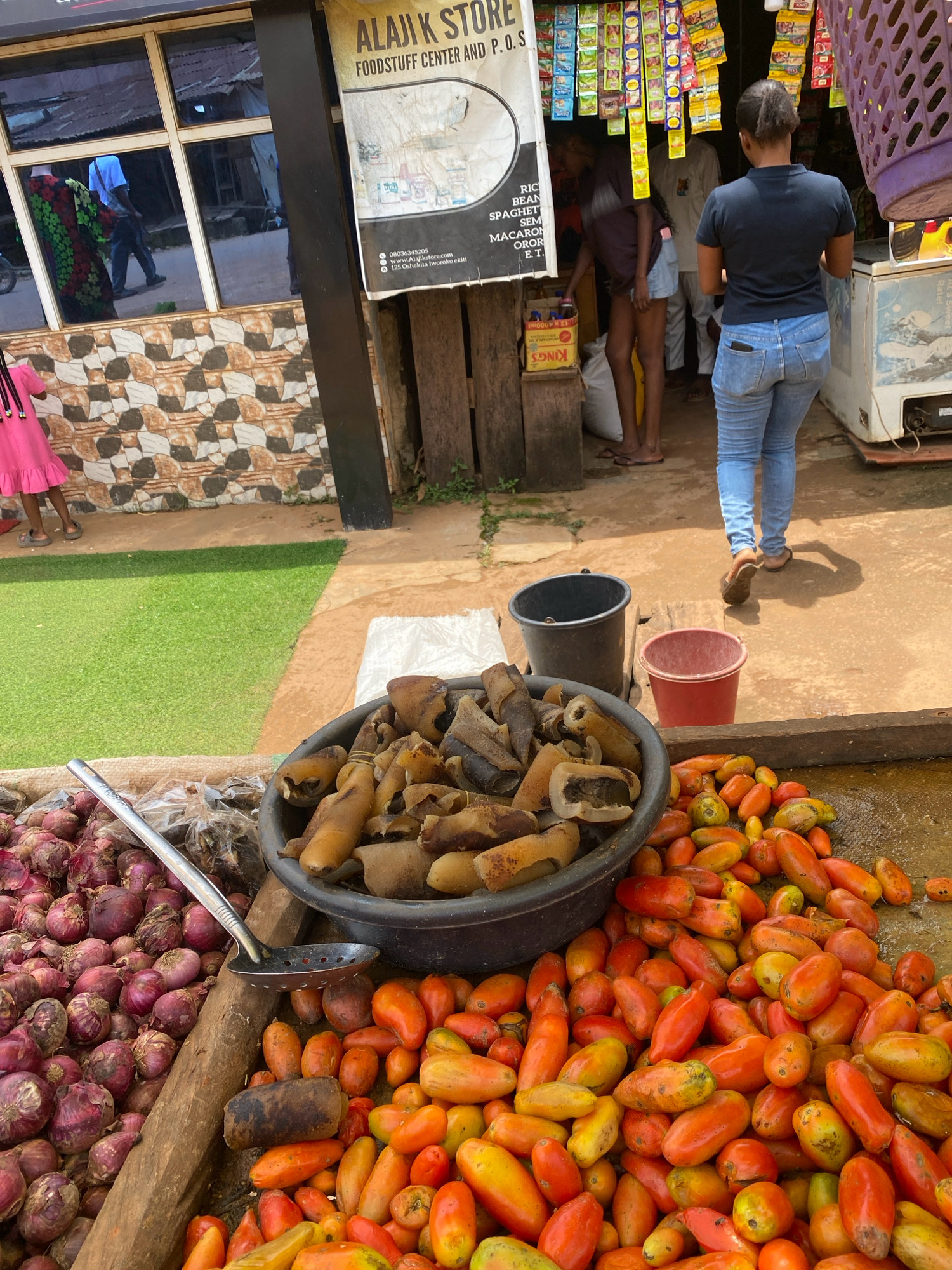 A street-level outdoor view shows a produce stall in front of a store. The store has a tiled facade and a sign that reads "ALAJIK STORE FOODSTUFF CENTER AND P.O.S." and advertises a phone number and website. A woman wearing a dark blue t-shirt and blue jeans walks away from the camera towards the store entrance. A young girl in a pink dress is standing on the left side of the frame. The produce stall displays a large pile of red onions, a black bowl filled with what appears to be dried meat or fish, and a large pile of red and orange tomatoes. Two buckets, one black and one red, are visible behind the produce. The ground is a mixture of dirt and concrete. The scene is set in the city of Iworoko, Nigeria.