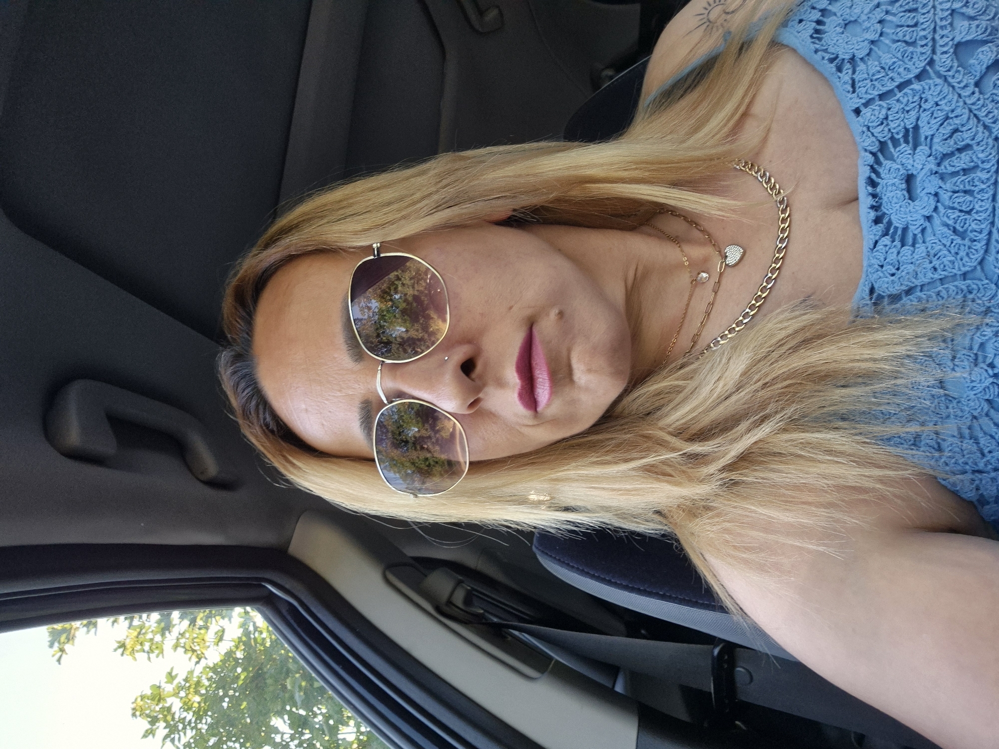 A medium shot captures a woman with blonde hair and sunglasses sitting in the driver's seat of a car. She is looking slightly to her right, with her lips parted and wearing pink lipstick. The sunglasses have gold rims and reflect trees and the sky. She is wearing a blue, crocheted top and two necklaces, one with a heart-shaped pendant. A seatbelt is visible across her shoulder. The interior of the car is dark, with a black headliner and a black seat. Outside the car window, green trees are visible against a bright sky.