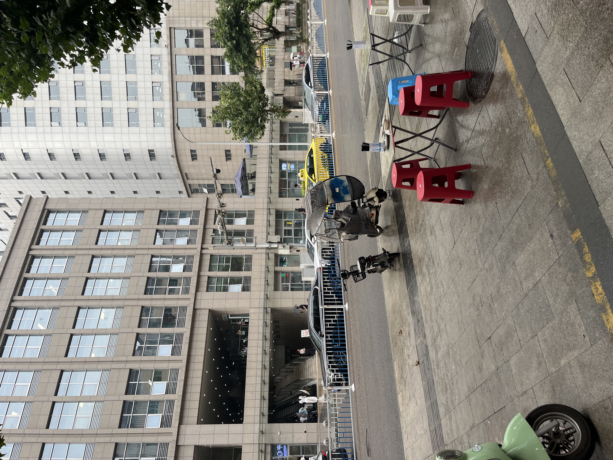 In Yuzhong District, China, an outdoor scene unfolds with a focus on a sidewalk café setup in front of modern buildings. The pavement is wet, suggesting recent rain, and marked with yellow and grey lines. Several red plastic stools surround two small metal tables. On one table, a metal bowl, a small dish with food, and a bottle of disinfectant are visible, along with chopsticks in a holder. A blue plastic stool is positioned at the other table, which also has white plastic stools nearby. To the left, a light green scooter is parked on the sidewalk, with its front wheel and fender prominently featured. Further back, a three-wheeled vehicle, possibly an electric scooter with a covered passenger area, is parked. Across the street, a black sedan with blue wheel accents is parked, preceded by a silver car and followed by a yellow taxi. Two white barriers with blue accents line the street. In the background, the lower floors of a large, modern building complex are visible, with a covered entrance featuring escalators and a few people standing. A taller, lighter-colored building stands to the right. Trees with green foliage are scattered throughout the scene, with some branches extending into the frame.