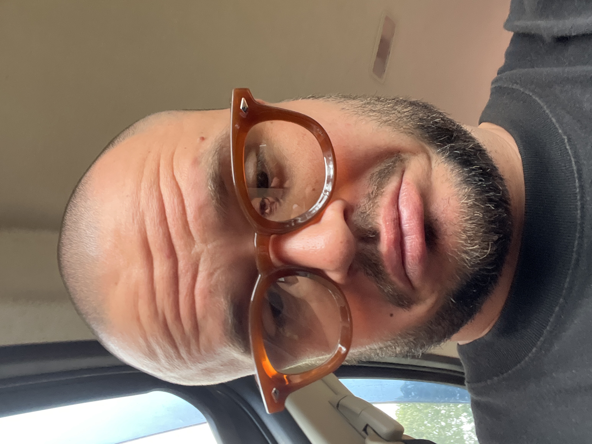 A close-up, frontal, eye-level, medium shot shows a bald, white man with a beard and brown-rimmed glasses, making a pouty face. The man is wearing a black t-shirt. He is sitting in the passenger seat of a vehicle. The background is blurred and shows a window with a view of trees. The interior of the car is visible, with a light-colored door panel and a dark-colored dashboard.