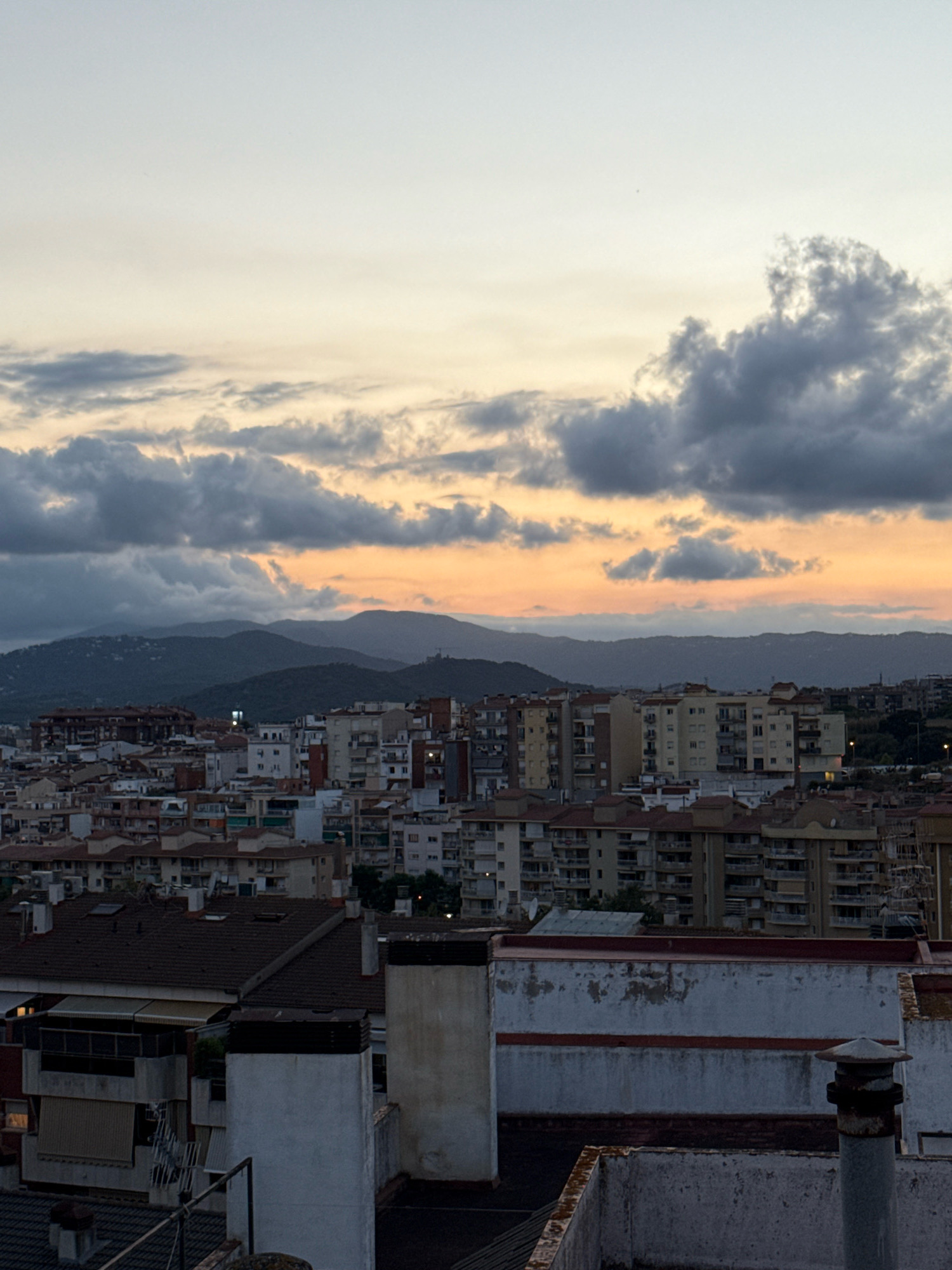 The image depicts a dusky sky over the city of Blanes, Spain. The sky is filled with dramatic clouds, tinged with the warm oranges and yellows of a sunset. In the foreground, the rooftops of residential buildings are visible, with various architectural styles and roof materials, including tiled and flat roofs. Chimneys and ventilation shafts are prominent features on many of the buildings. Beyond the immediate rooftops, a dense urban landscape stretches out, with numerous apartment buildings of varying heights and colors. In the distance, a range of rolling, tree-covered hills forms a natural backdrop to the city. The lighting suggests it is either early morning or late evening, with shadows cast by the buildings and the low-lying sun. There are no visible people or discernible actions taking place within the frame.