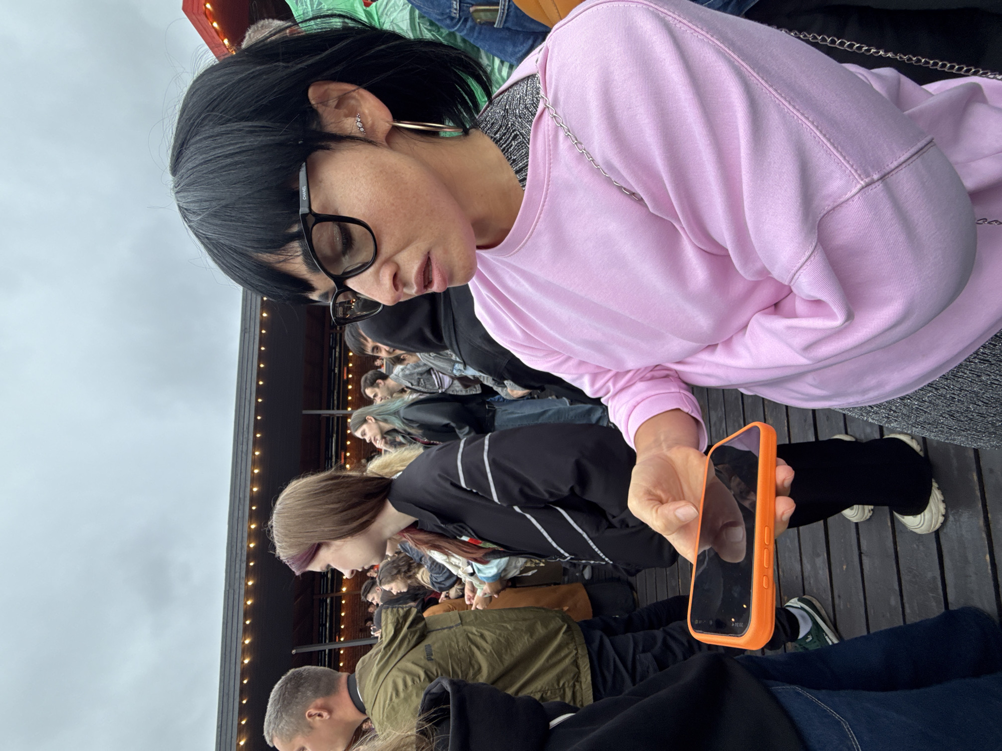 The media file shows a group of people standing or sitting on a wooden deck, with a blurred background of what appears to be a building with lights. The sky is overcast, suggesting a cloudy day. The people are dressed in casual clothing.
The main subject in the foreground is a woman with dark hair, wearing glasses and a pink sweatshirt. She is holding an orange smartphone in her right hand and looking down at it. Her expression is neutral. Another person, possibly a woman with brown hair, is visible to her left, also looking down. Further back, other individuals are seen, some looking at their phones, others in conversation.
The scene appears to be an outdoor gathering or event. The wooden deck and the strung lights hint at a casual, possibly festive atmosphere, like a concert, market, or outdoor dining area. The overcast sky and the clothing suggest a temperate climate.
There is no visible text in the media file, except for what might be on the screen of the phone, which is too blurred to read. The overall mood seems relaxed and informal. The location context provided is Saint Petersburg, Russia, which fits the possibility of an outdoor event in a city.