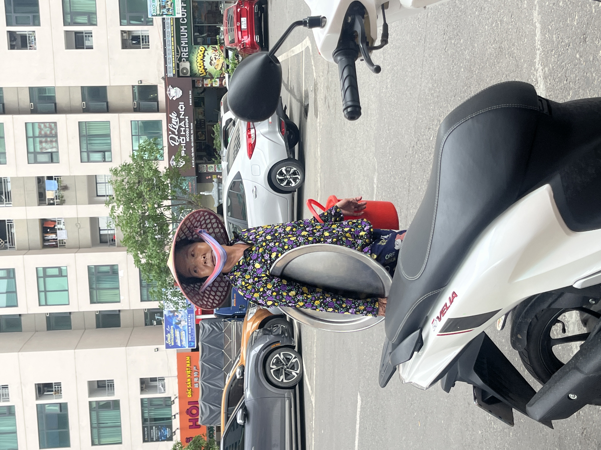 An elderly Vietnamese woman, wearing a traditional conical hat (nón lá) and a purple long-sleeved shirt with a floral pattern, stands on a sidewalk in an urban setting. She holds a large, round silver tray in her left arm and has a red bucket nearby. Her face, partially obscured by a light-colored scarf, looks directly at the camera with a calm expression.
She is positioned next to a white scooter, branded "FOVILIA", which occupies the right foreground of the image. The setting appears to be a busy street in Bac Nha Trang Ward, Vietnam, under bright, clear daylight. In the background, several multi-story buildings with numerous windows and balconies line the street. Parked cars, including a white hatchback, a red sedan, and a dark grey SUV, are visible on the road.
Various storefront signs are visible on the ground floors of the buildings. These include "D'Linh PHỞ HÀ NỘI NOODLE SOUPS" with "ĐỒNG, NHA TRANG" below, suggesting a noodle restaurant. Other signs advertise "PREMIUM COFFEE," "LOCOMOCO," "HỘI ĐẶC SẢN VIỆT NAM" (Specialties of Vietnam Association), and "VIET NAM DENTAL" accompanied by "Smile WITH EXPERTS". Another sign shows "FASHION" and a partial "ORF." A green tree stands between the buildings and the street, adding natural elements to the urban landscape.