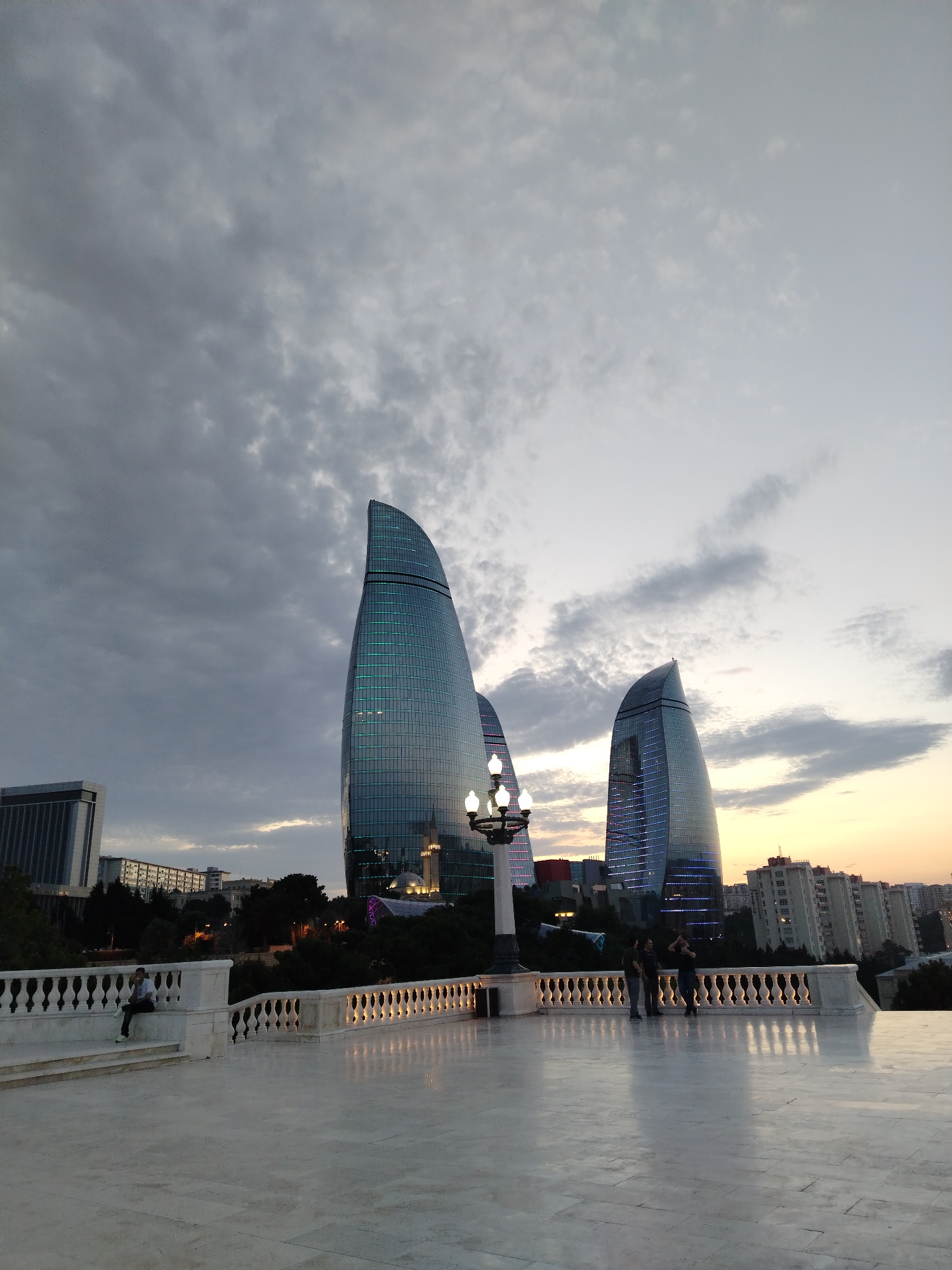 The image captures a twilight scene in Sabail Raion, Azerbaijan, featuring the distinctive Flame Towers. In the foreground, a white marble terrace with a decorative balustrade overlooks a cityscape. Several people are visible on the terrace, with one individual sitting on the steps to the left and a group of three standing near the balustrade to the right. The Flame Towers, illuminated by blue lights, dominate the background, with other city buildings and trees visible between them and the foreground terrace. The sky is cloudy, with hints of sunset colors near the horizon. The lighting suggests it is either dusk or early evening, with artificial lights beginning to punctuate the urban landscape.