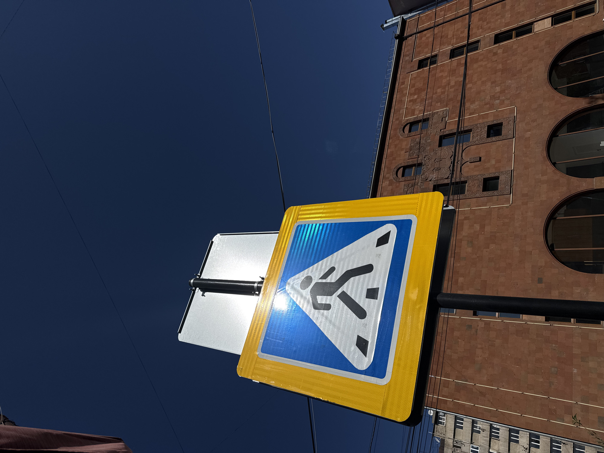 The image captures a low-angle perspective looking upwards at a street sign, a building, and a deep blue sky in Yerevan, Armenia.
In the foreground, a reflective pedestrian crossing sign is prominently displayed. It features a bright yellow outer border, enclosing a blue triangular area with a white border. Inside the triangle, a black silhouette of a person walking is visible. Strong sunlight creates a brilliant glare across the sign's surface, particularly on the blue and white sections. The sign is attached to a black pole, and behind it, a plain silver-backed sign is partially visible.
To the right, a tall, multi-story building with reddish-brown brick or stone cladding occupies the upper portion of the frame. Its distinct architecture includes several large circular windows on the right side and numerous rectangular windows. The building's facade is adorned with intricate carved relief patterns, notably a large, ornate panel with detailed motifs in its central section.
The background is dominated by a clear, deep blue sky, indicating bright, sunny weather. Several thin black electrical or utility wires crisscross the sky, passing both in front of the building and the signs. The intense sunlight suggests it is daytime. No legible text is visible on the sign or the building's facade.