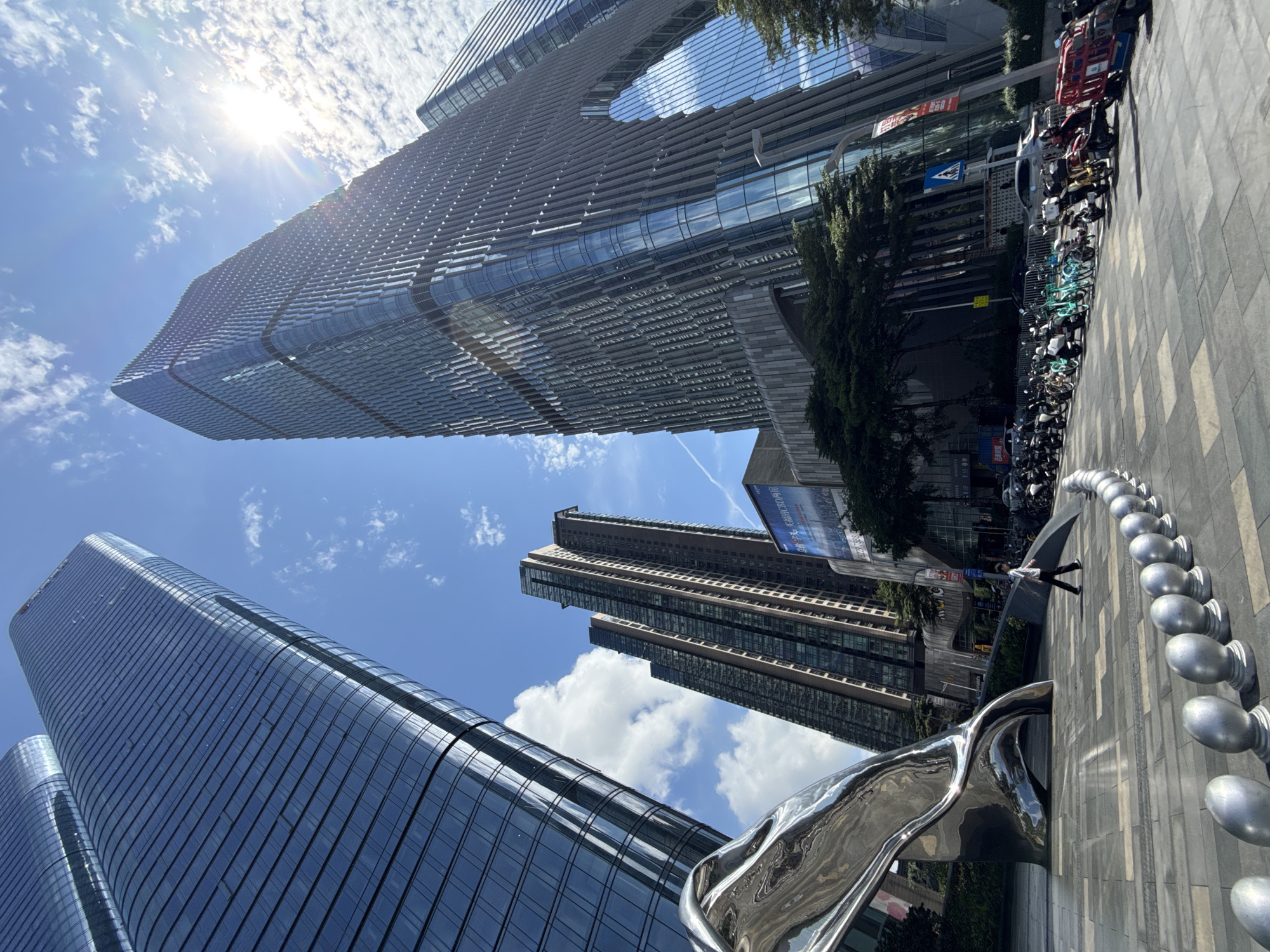 The image captures a low-angle view in the Futian District of China, showcasing modern architecture against a bright, partly cloudy sky. Prominent are several skyscrapers, their glass exteriors reflecting the sunlight and sky. The building in the upper center features a distinctive design with a large horizontal opening.
On the ground level, there is a contemporary art installation, possibly made of polished metal, reflecting the surrounding buildings. A row of metallic orbs follows the edge of the sidewalk. Parked bicycles and scooters line the curb. A person is seen walking in the mid-ground. The environment suggests a bustling urban setting.