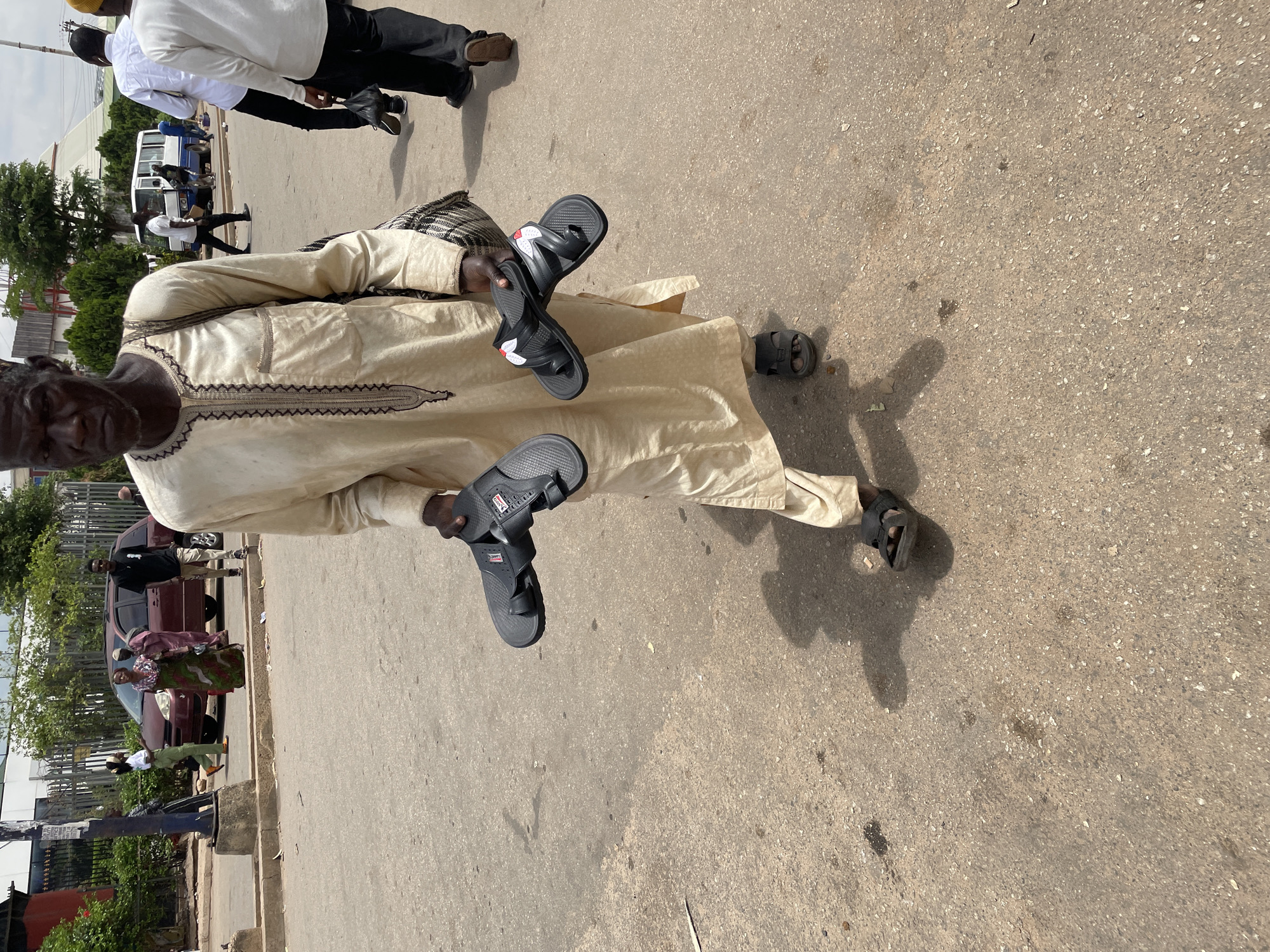 An elderly man in Jos, Nigeria, walks along a paved road, holding two pairs of sandals in his hands. He is wearing a long, light-colored robe with embroidered detail around the neckline, and sandals on his feet. Other people are walking in the background, and cars and buildings are visible. The road appears to be in a busy area of the city.