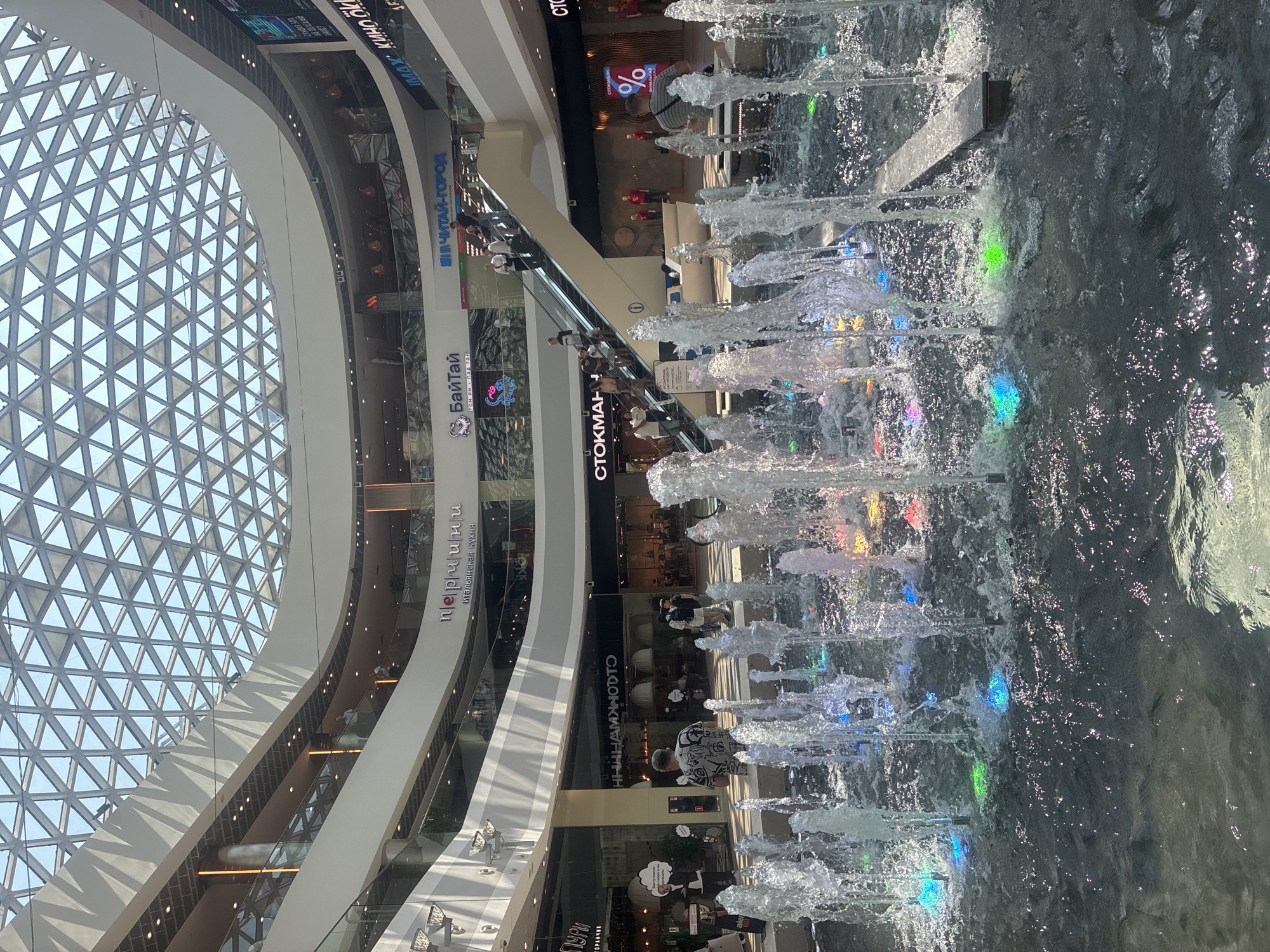 The image shows the interior of a multi-level shopping mall with a large, geodesic dome-shaped skylight. In the foreground, there is a water fountain with illuminated jets, casting colorful reflections on the water's surface. Several people are visible on different levels of the mall, some walking, others on an escalator. Shops and signage are present on the various floors. The overall environment appears to be a modern and well-lit commercial space. The signage visible in the image, specifically "МОРЕМОЛЛ" and "IMAX," indicates the location is the MoreMall shopping center in Sochi, Russia.