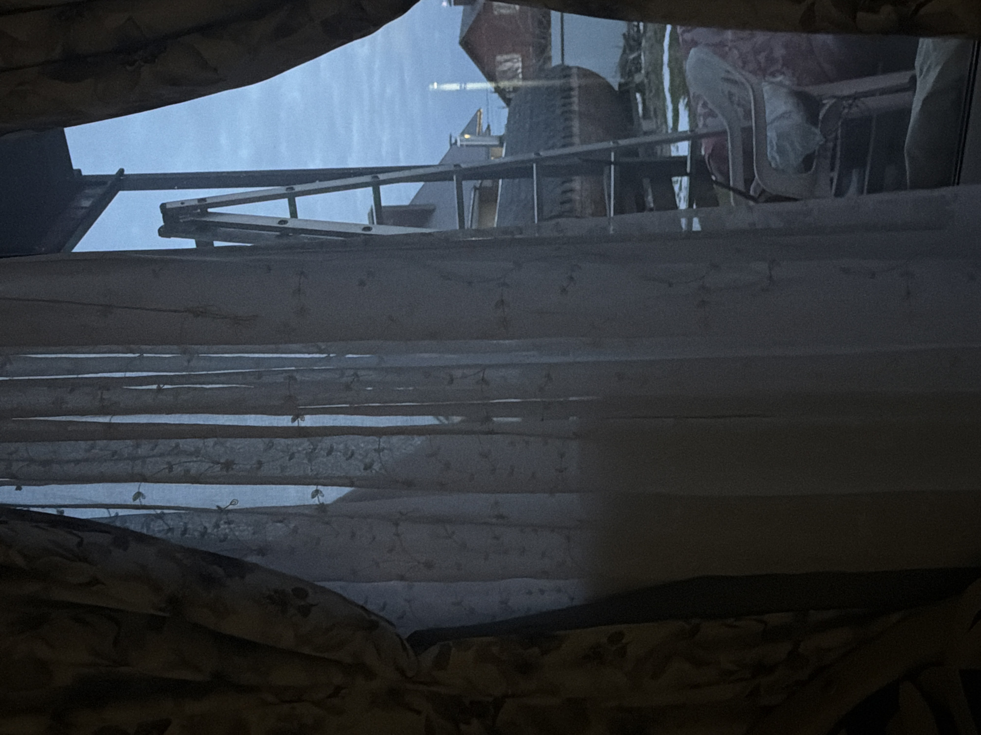 View through a window in Bol'shaya Shil'na, Russia. The window is partially obscured by a floral-patterned curtain. Outside the window, a building with a red roof and a stone structure is visible. A ladder is leaning against the building. A balcony with furniture is also visible outside the window. The sky is overcast.